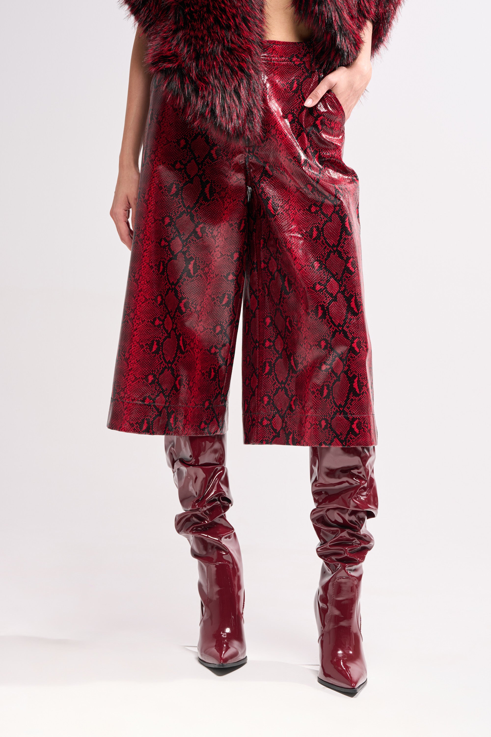 Model wearing the EXTRA IS MY MIDDLE NAME SNAKE BERMUDA SHORT, paired with matching shiny, red, embossed snakeskin boots and a fur-trimmed jacket, hand in pocket, set against a plain background.