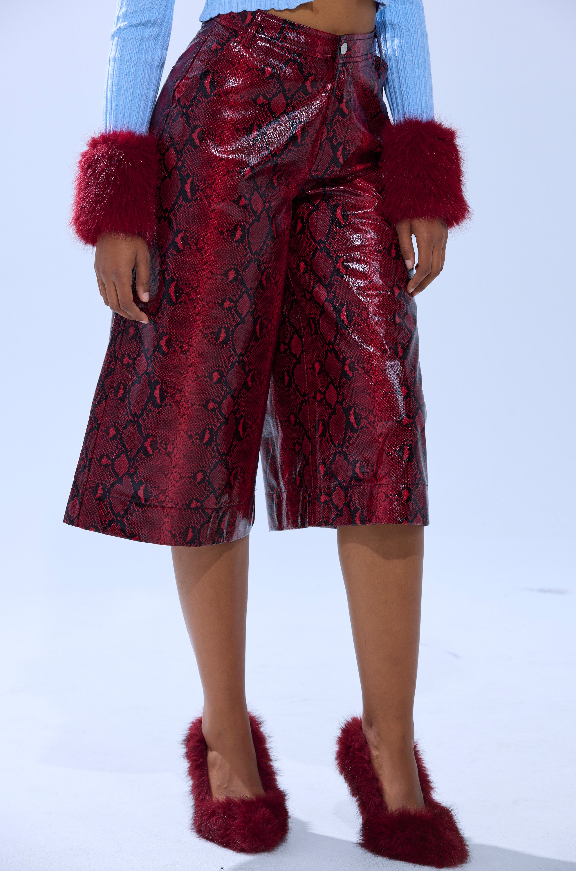 Wearing the EXTRA IS MY MIDDLE NAME SNAKE BERMUDA SHORT in shiny red embossed snakeskin, a light blue ribbed top with red furry cuffs, and matching red furry heeled shoes, this person stands against a plain light background.