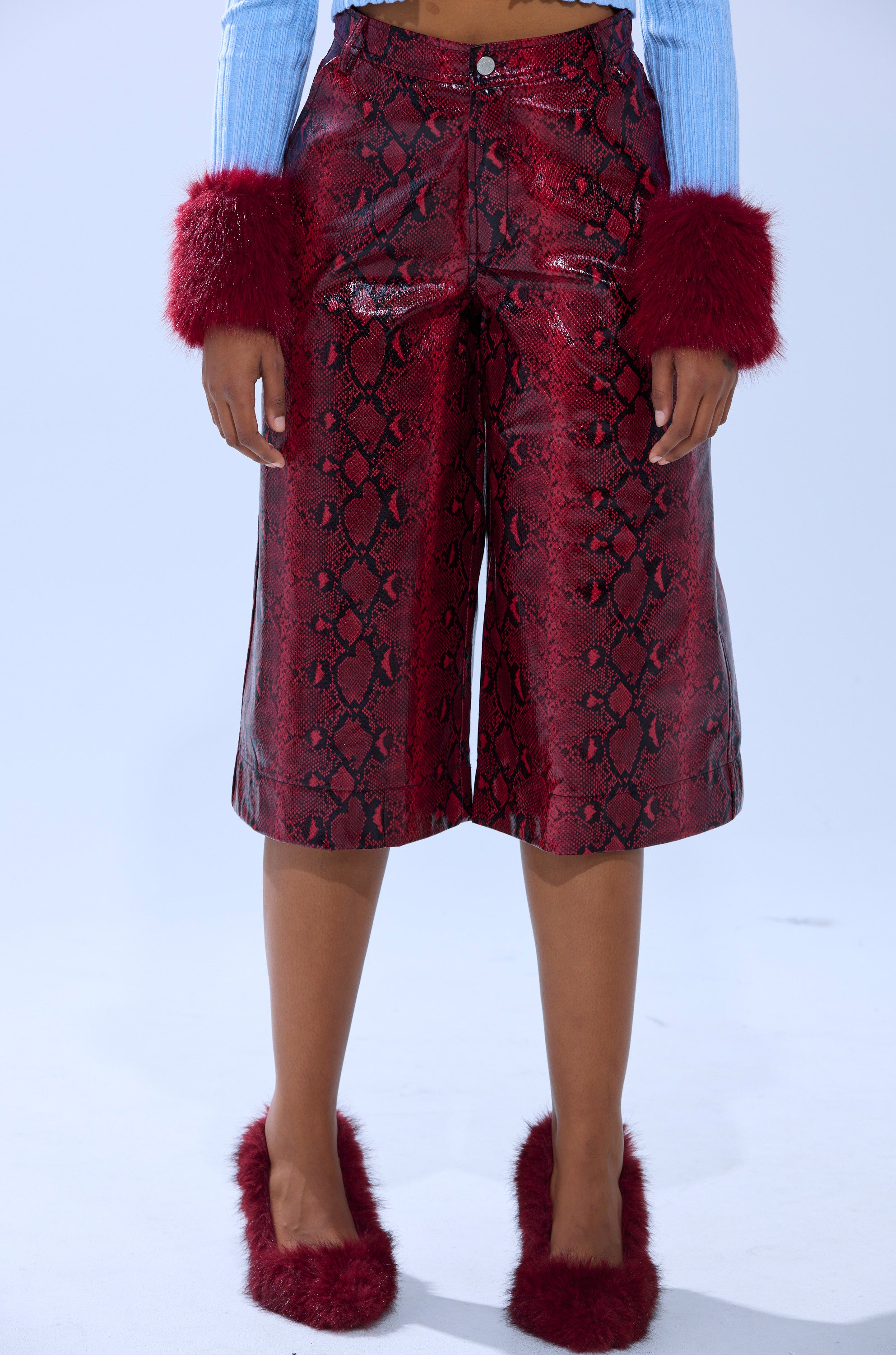 Wearing EXTRA IS MY MIDDLE NAME SNAKE BERMUDA SHORTS in red snakeskin print, paired with a light blue ribbed top with red fuzzy cuffs and matching red fuzzy slippers. Only the lower half is visible against a plain light background.