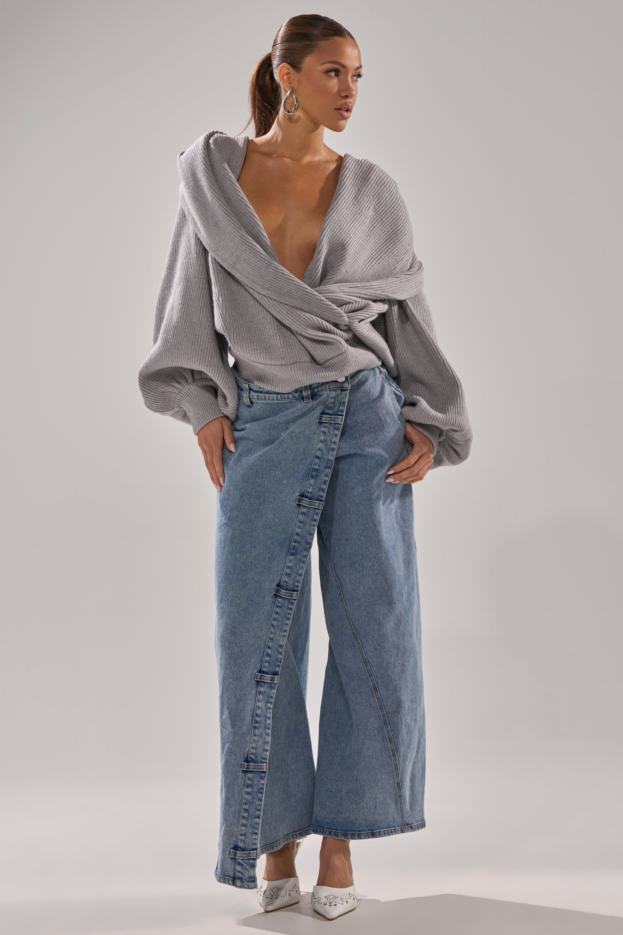 A woman models the COVERED UP RELAXED DENIM PANT, a wide-leg, wrap-style denim, paired with a loose off-shoulder gray sweater and pointed white heels. She accessorizes with hoop earrings and a high ponytail against a plain gray background.