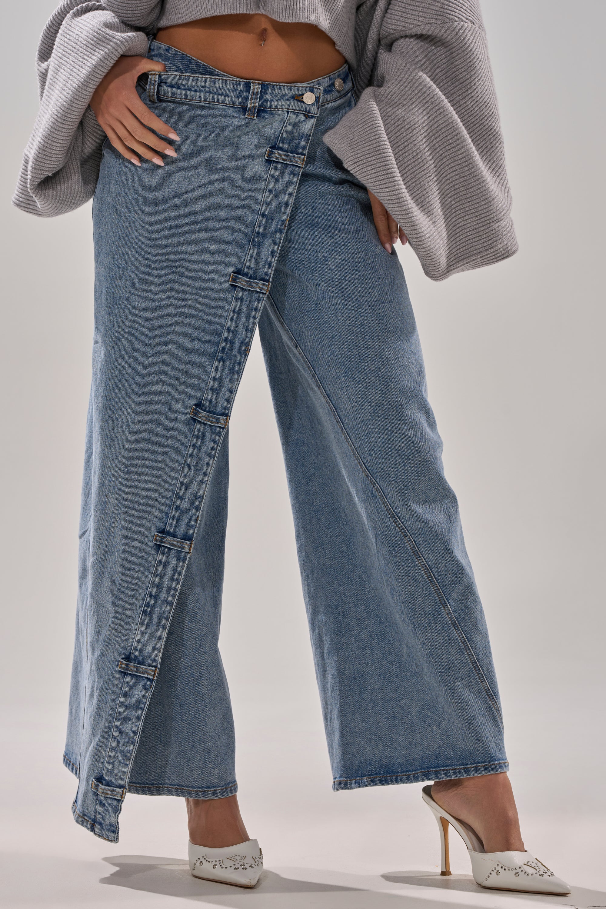 Woman wears COVERED UP RELAXED DENIM PANT—high-rise, wide-leg blue jeans with an asymmetrical front panel and buttons—styled with a gray sweater and white pointed-toe heeled mules for a modern, relaxed look.