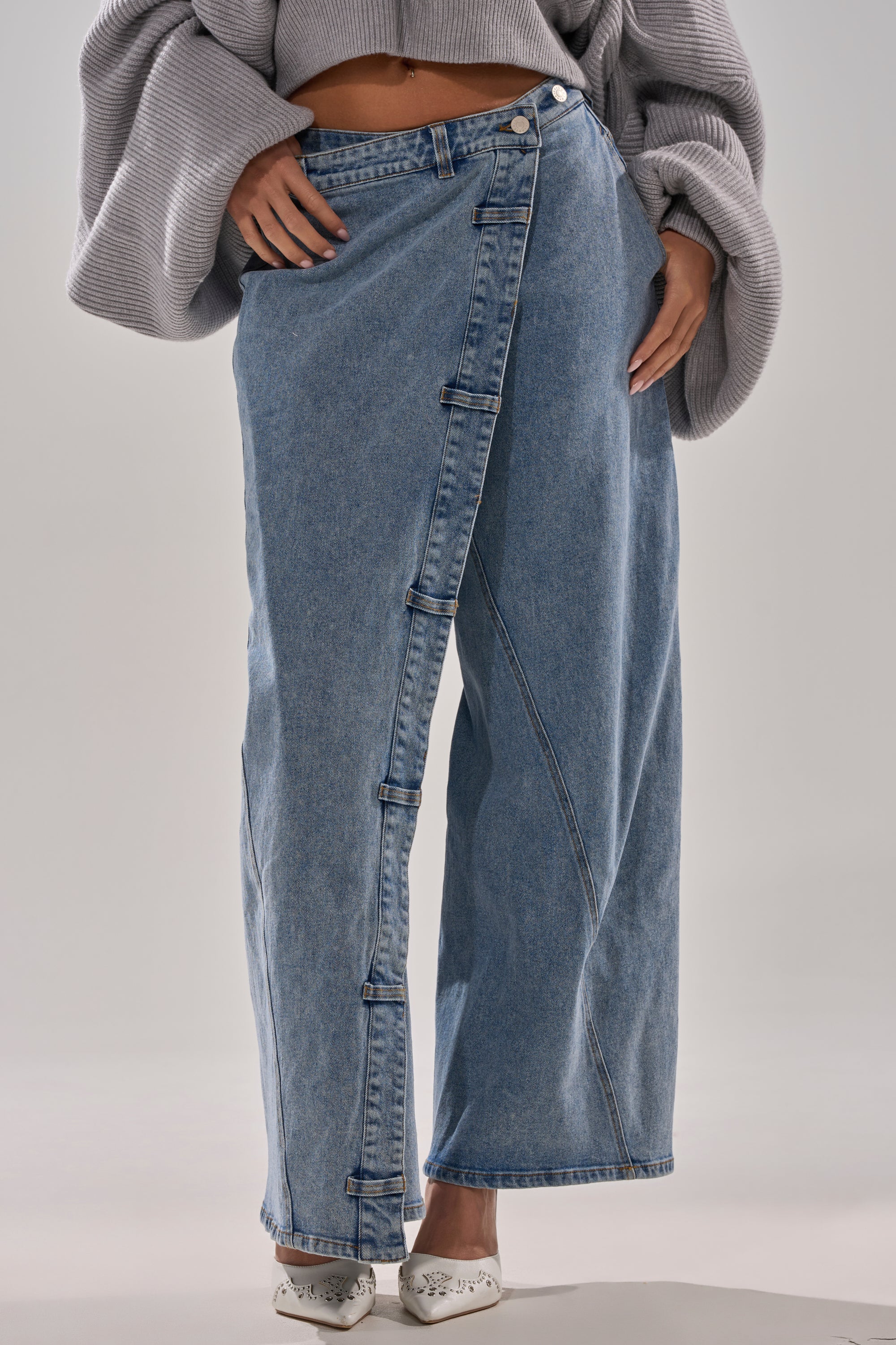 A person wearing COVERED UP RELAXED DENIM PANT featuring a high-waisted, relaxed fit and asymmetric button detail, paired with a cropped oversized grey sweater; only the lower torso and legs are visible.