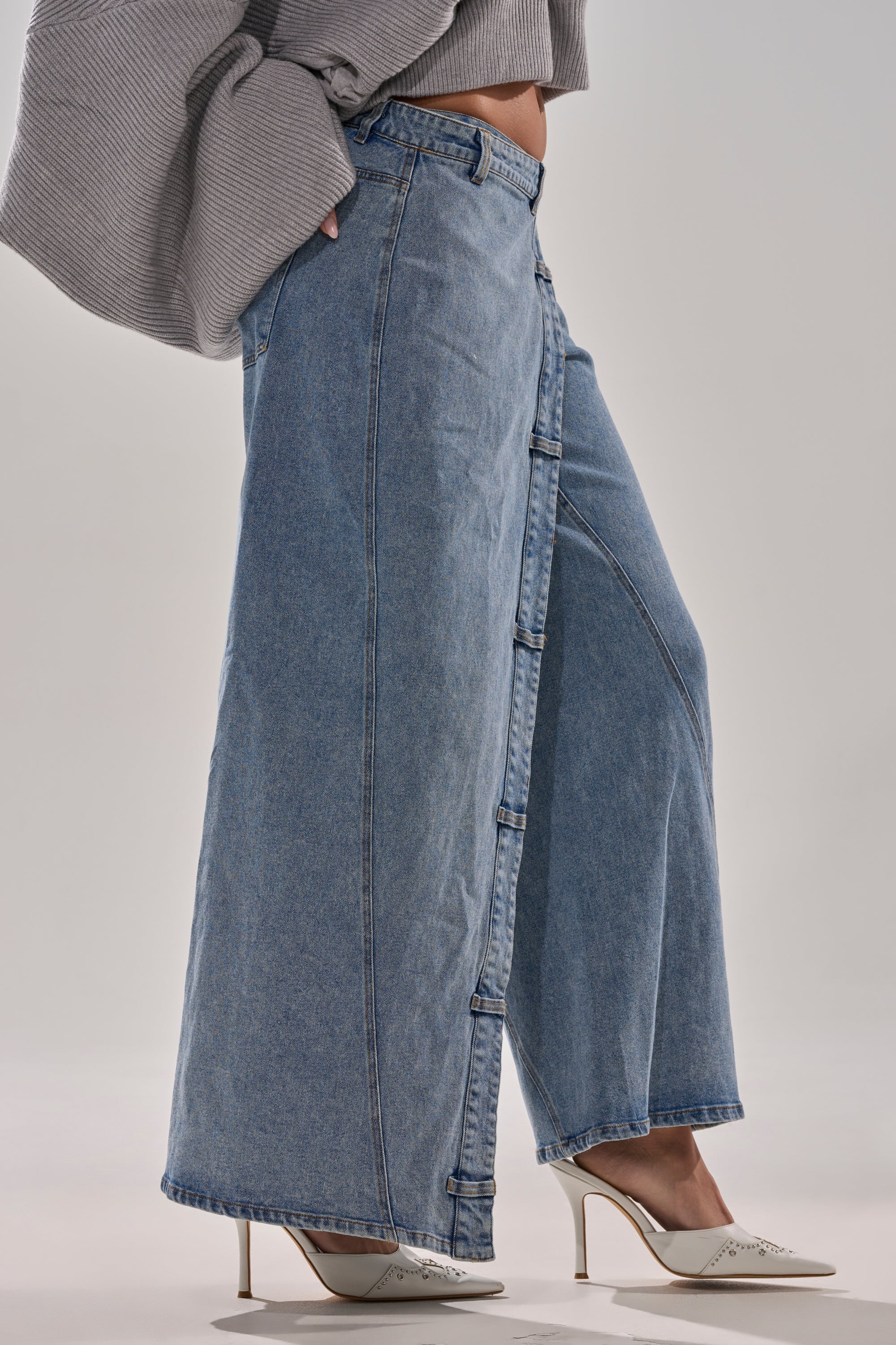 Wearing the COVERED UP RELAXED DENIM PANT with side buttons, a loose gray sweater, and white heeled shoes, this look creates a chic maxi skirt illusion against a plain background.