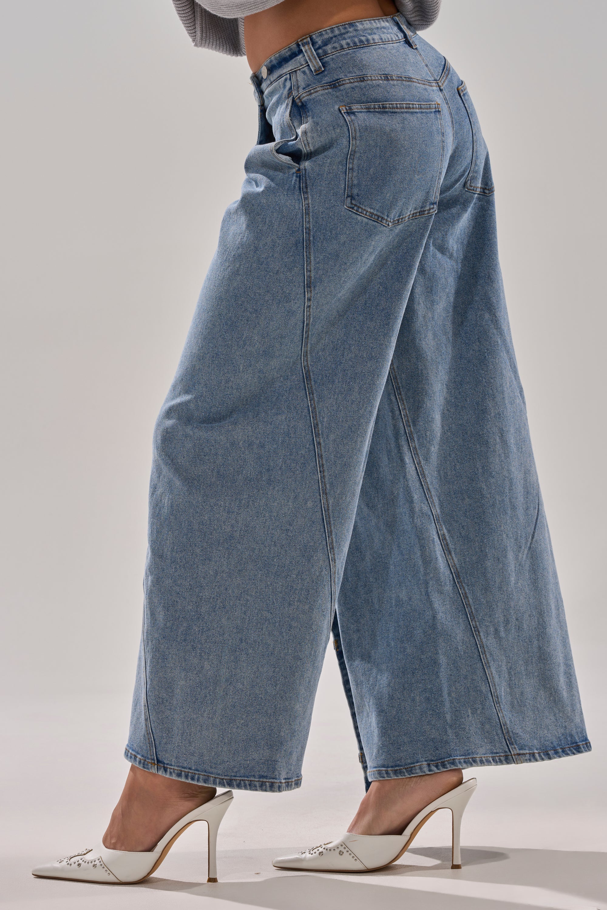 A person models the COVERED UP RELAXED DENIM PANT, featuring a wide-leg, high-rise fit, styled with white pointed-toe heels against a simple light background.