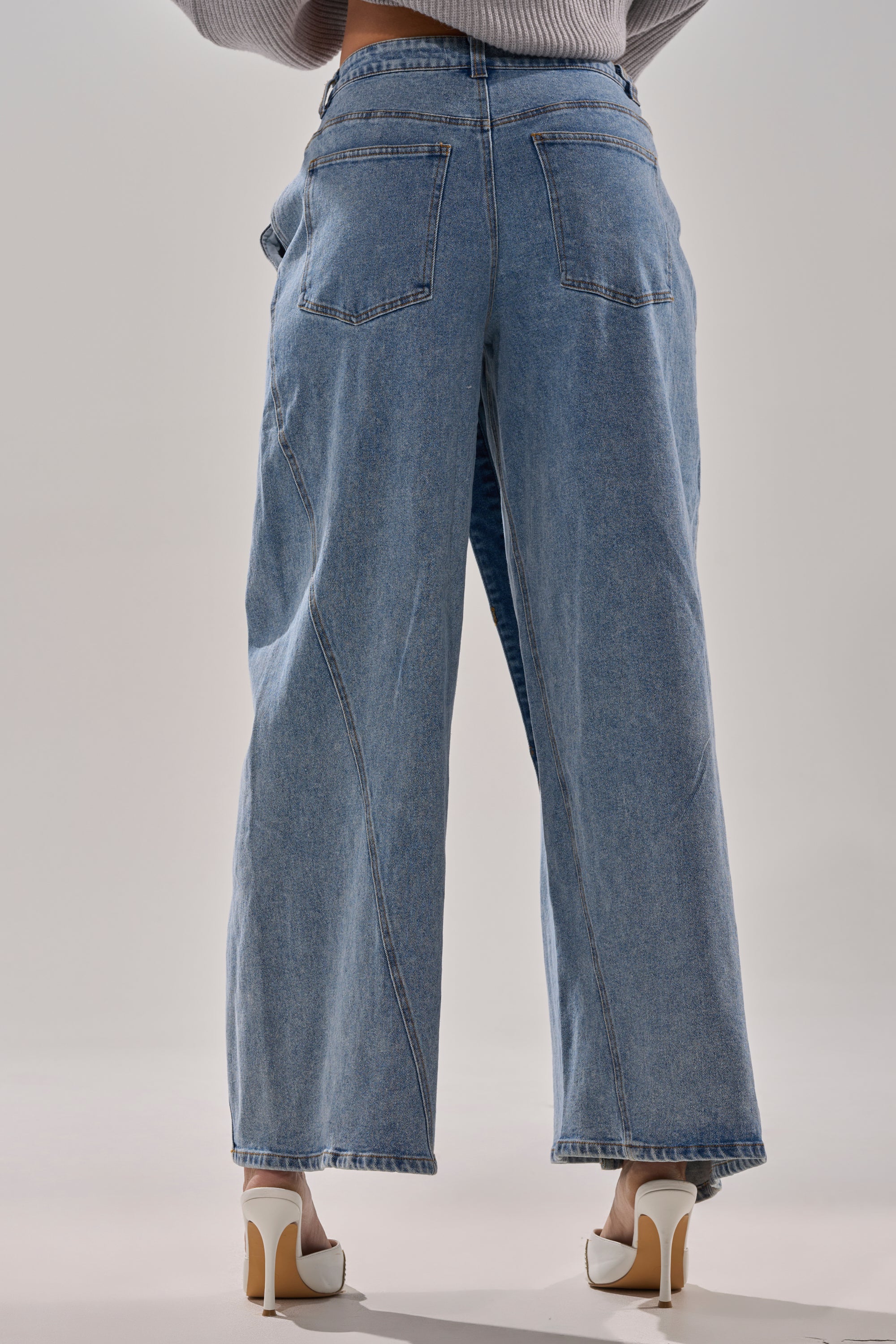 A person models the COVERED UP RELAXED DENIM PANT, featuring a straight-leg fit, with white high heels. Posed with their back to the camera against a plain light backdrop, only their lower half is visible.