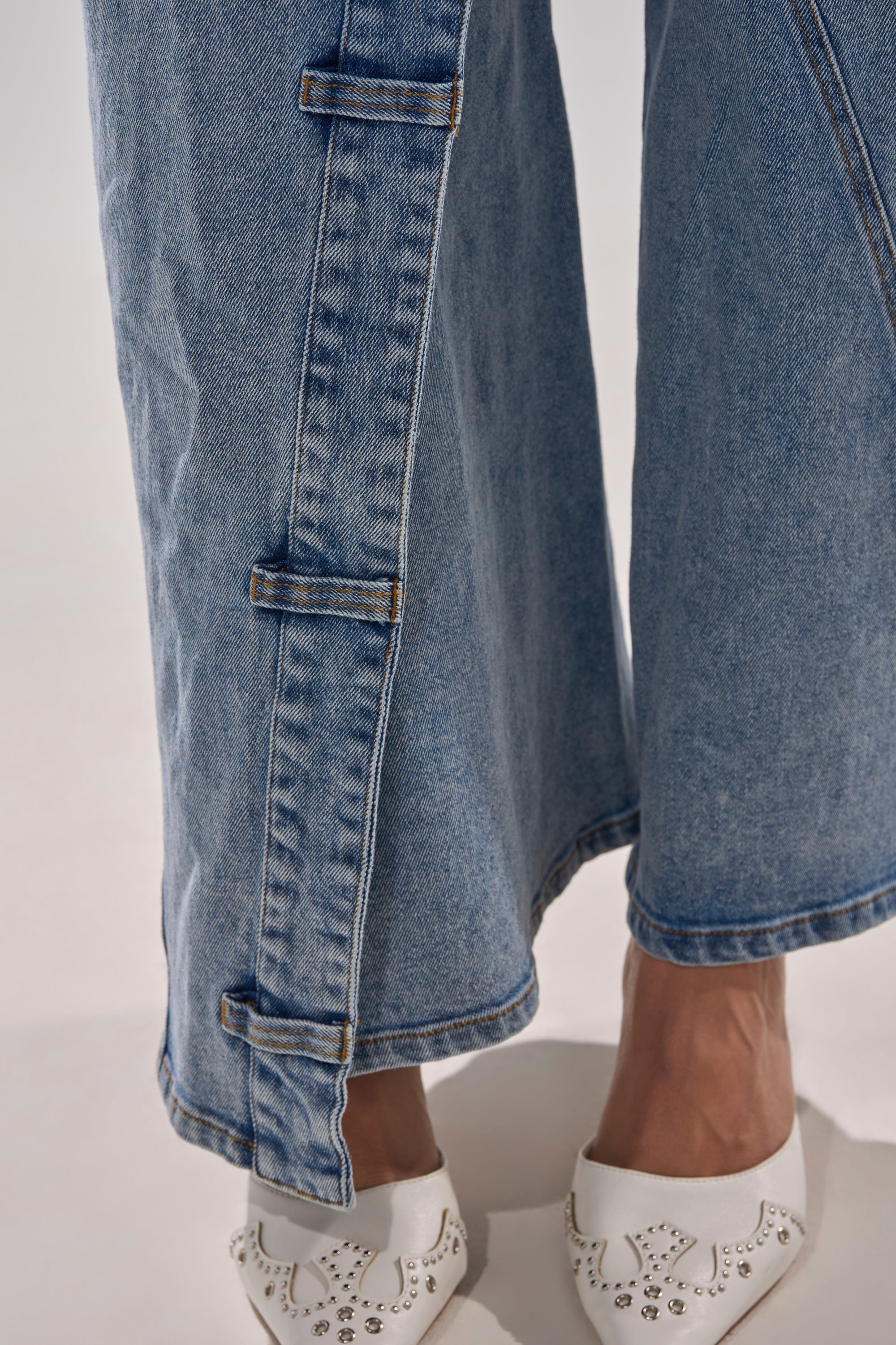 Close-up of a person wearing the COVERED UP RELAXED DENIM PANT, a high-rise blue wide-leg jean with three rectangular fabric loops on the side seam, paired with white cut-out flats accented by silver studs.