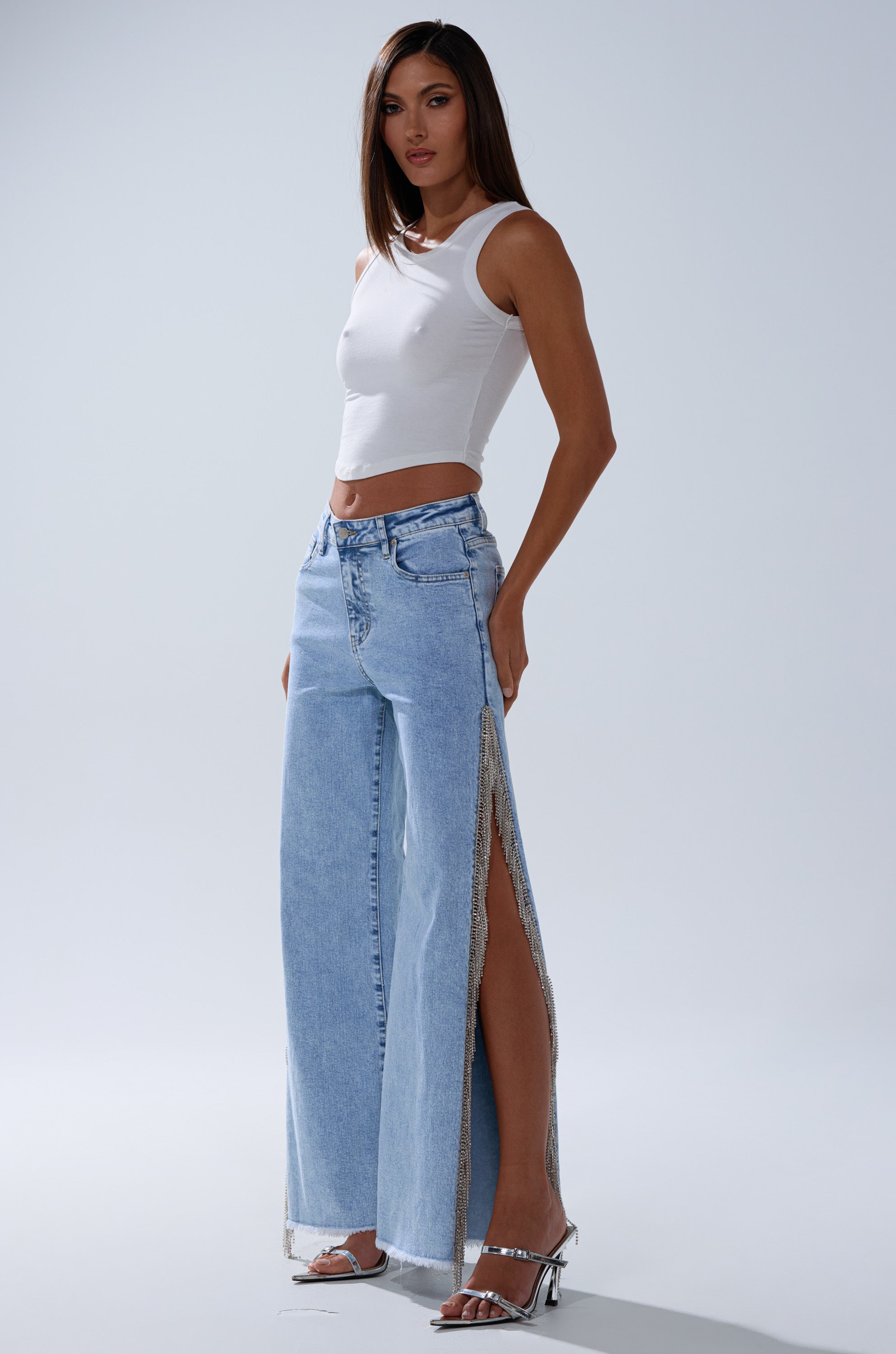 A woman poses against a plain light grey background wearing ESSENTIAL RELAXED FIT BLING DENIM JEANS with high side slits, a white sleeveless crop top, and silver high-heeled sandals.