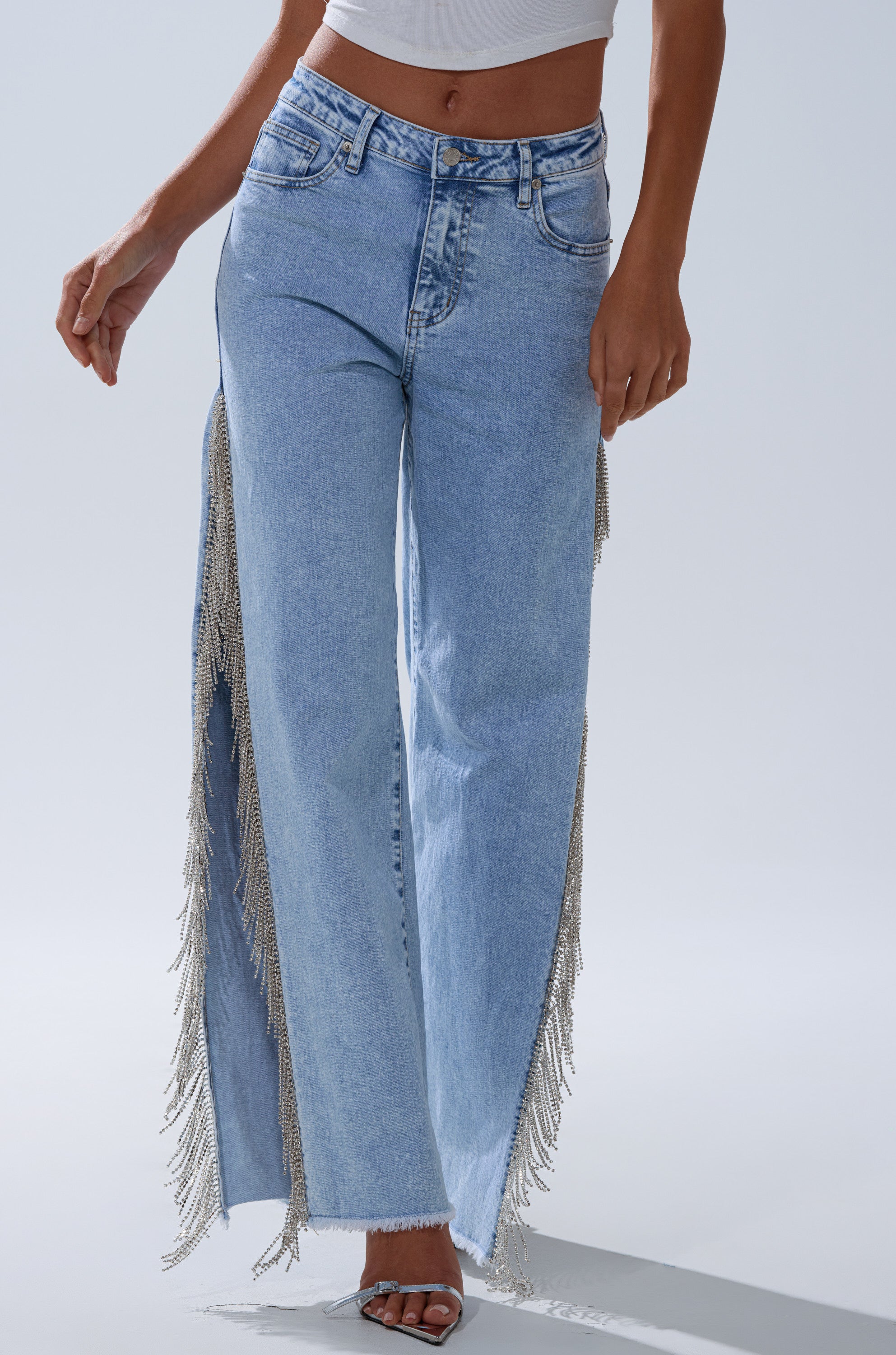 A person wears ESSENTIAL RELAXED FIT BLING DENIM JEANS featuring light blue denim and silver fringe trim along the side seams, styled with a white crop top and open-toe heels, standing against a plain light background.