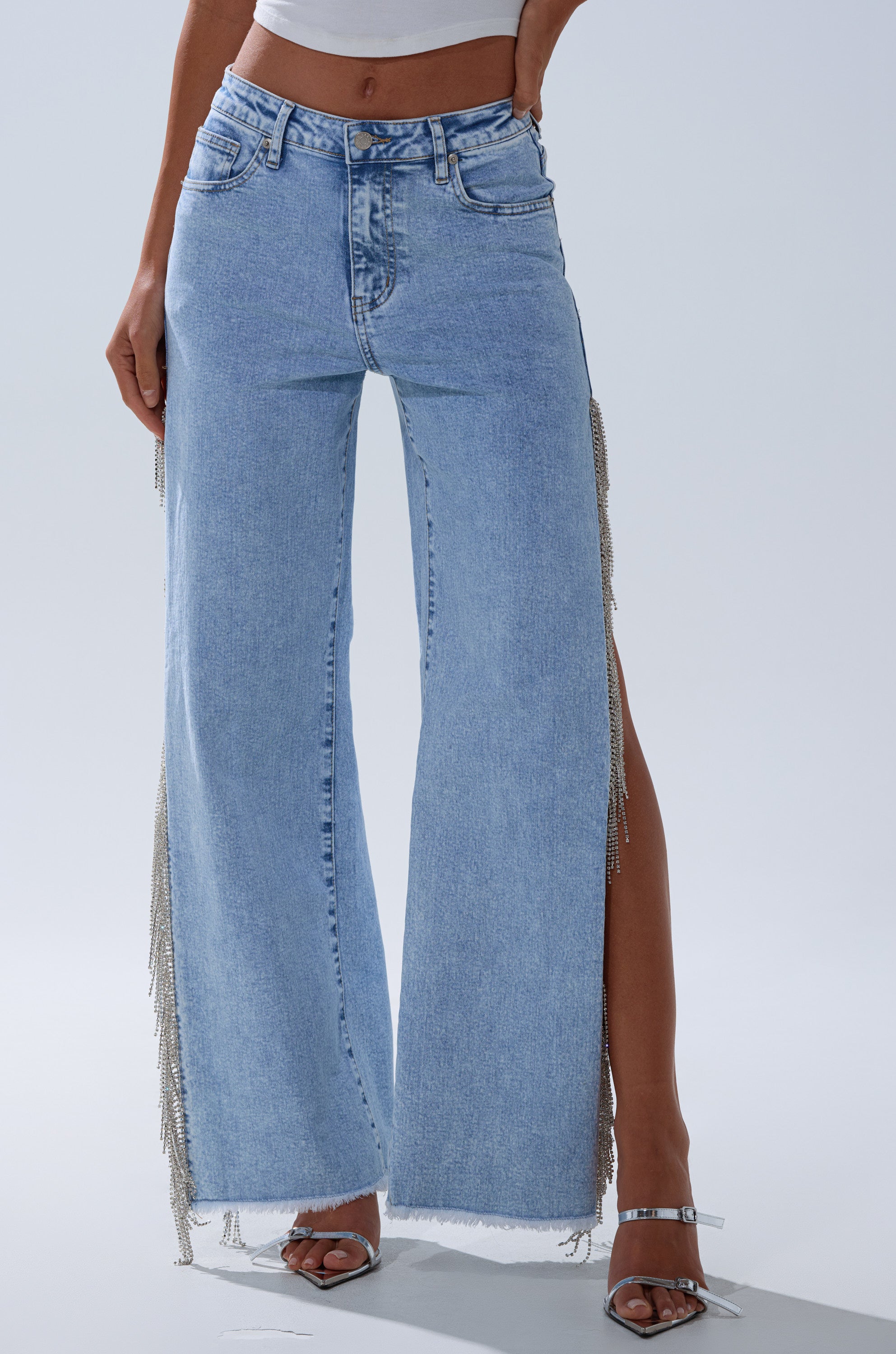 Someone wearing the ESSENTIAL RELAXED FIT BLING DENIM JEANS—light blue with rhinestone trim, wide legs, high side slits, and silver fringe—paired with a white crop top and silver high-heeled sandals.