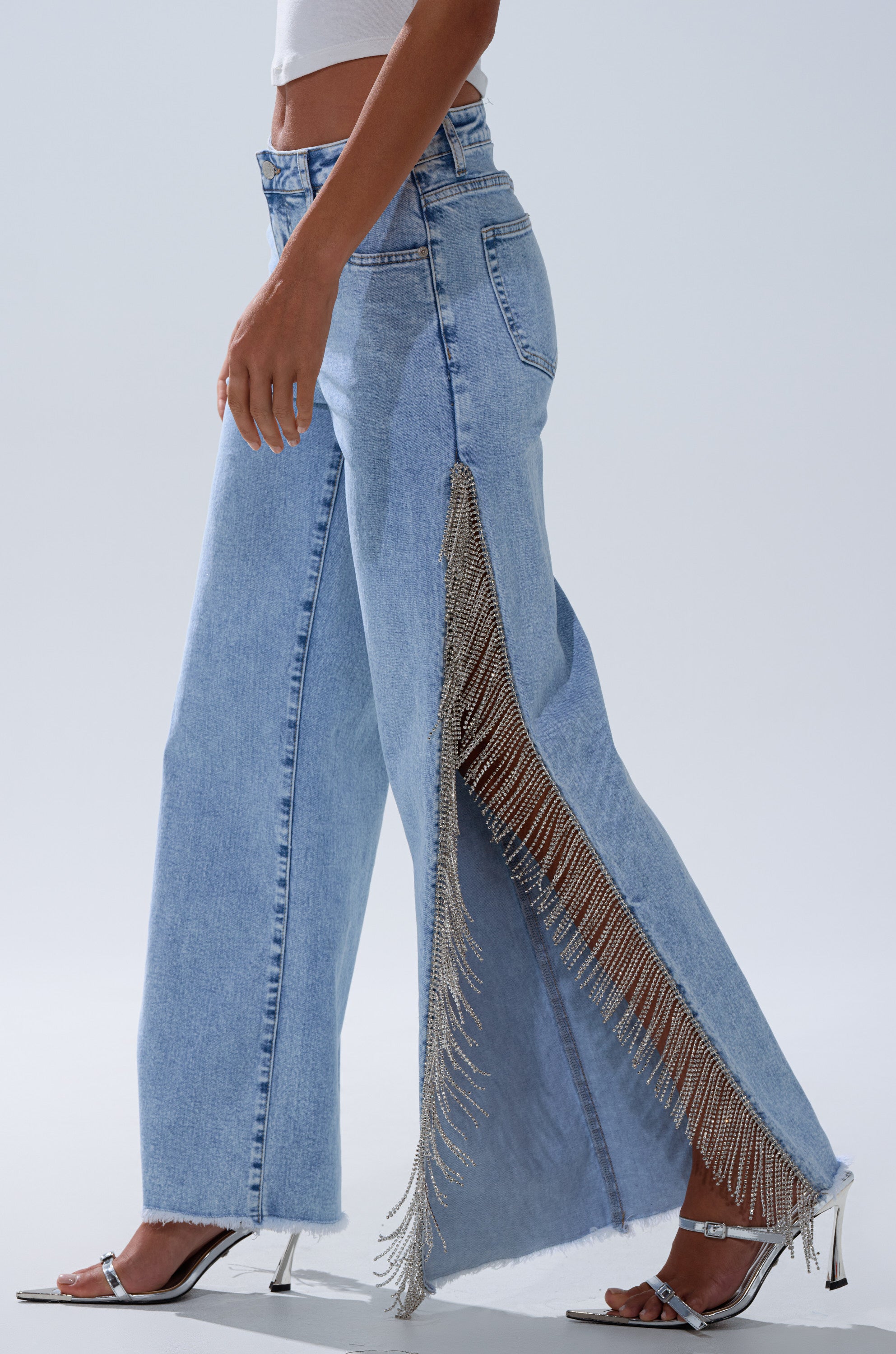 Wearing ESSENTIAL RELAXED FIT BLING DENIM JEANS with long silver fringe on a high side slit, paired with a white crop top and silver heeled sandals, this look stands out against a plain background.