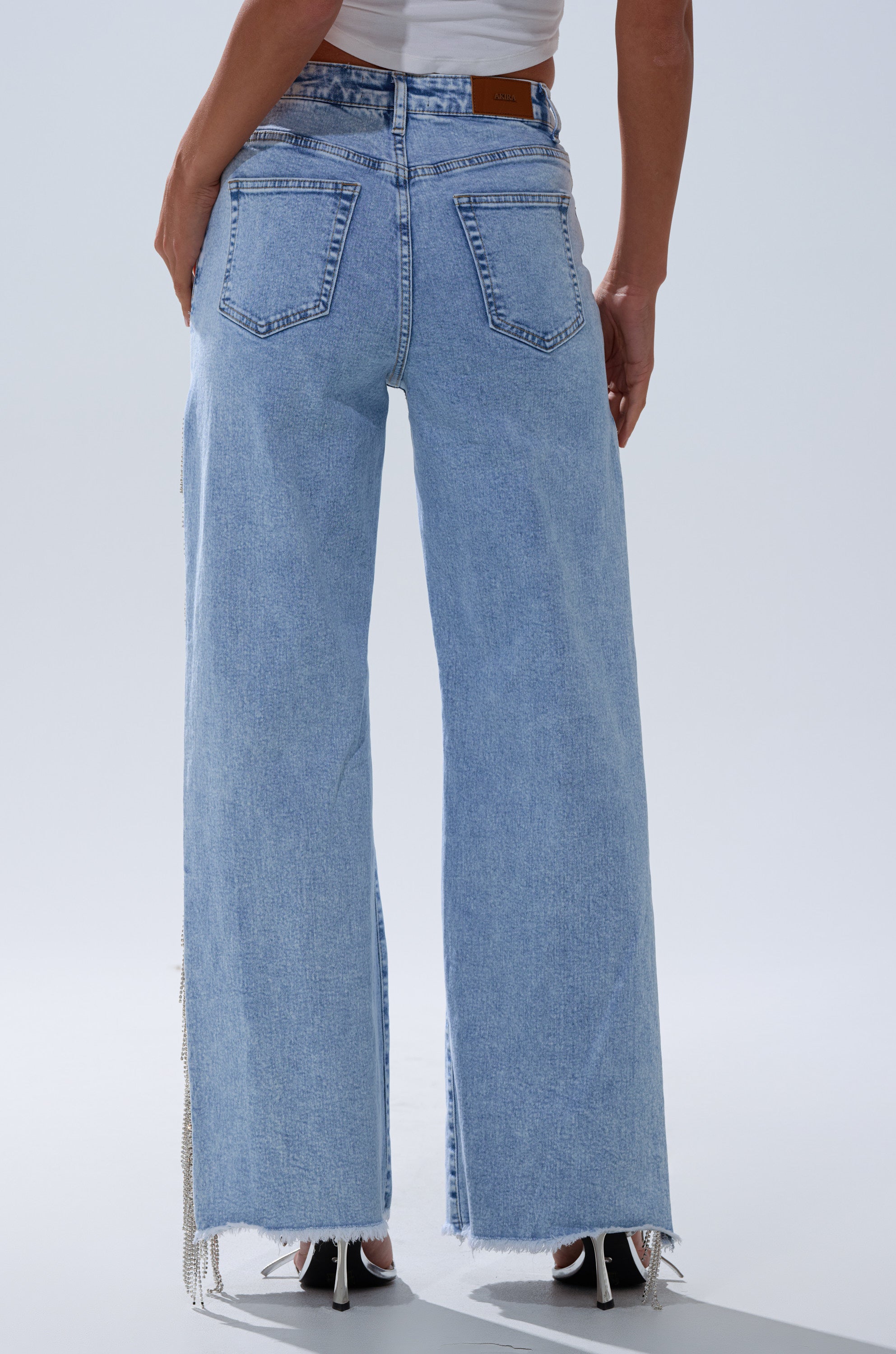 A person is shown from the back in light blue, high-waisted ESSENTIAL RELAXED FIT BLING DENIM JEANS with frayed hems, styled with a white sleeveless top and black heeled sandals for an effortlessly chic look.