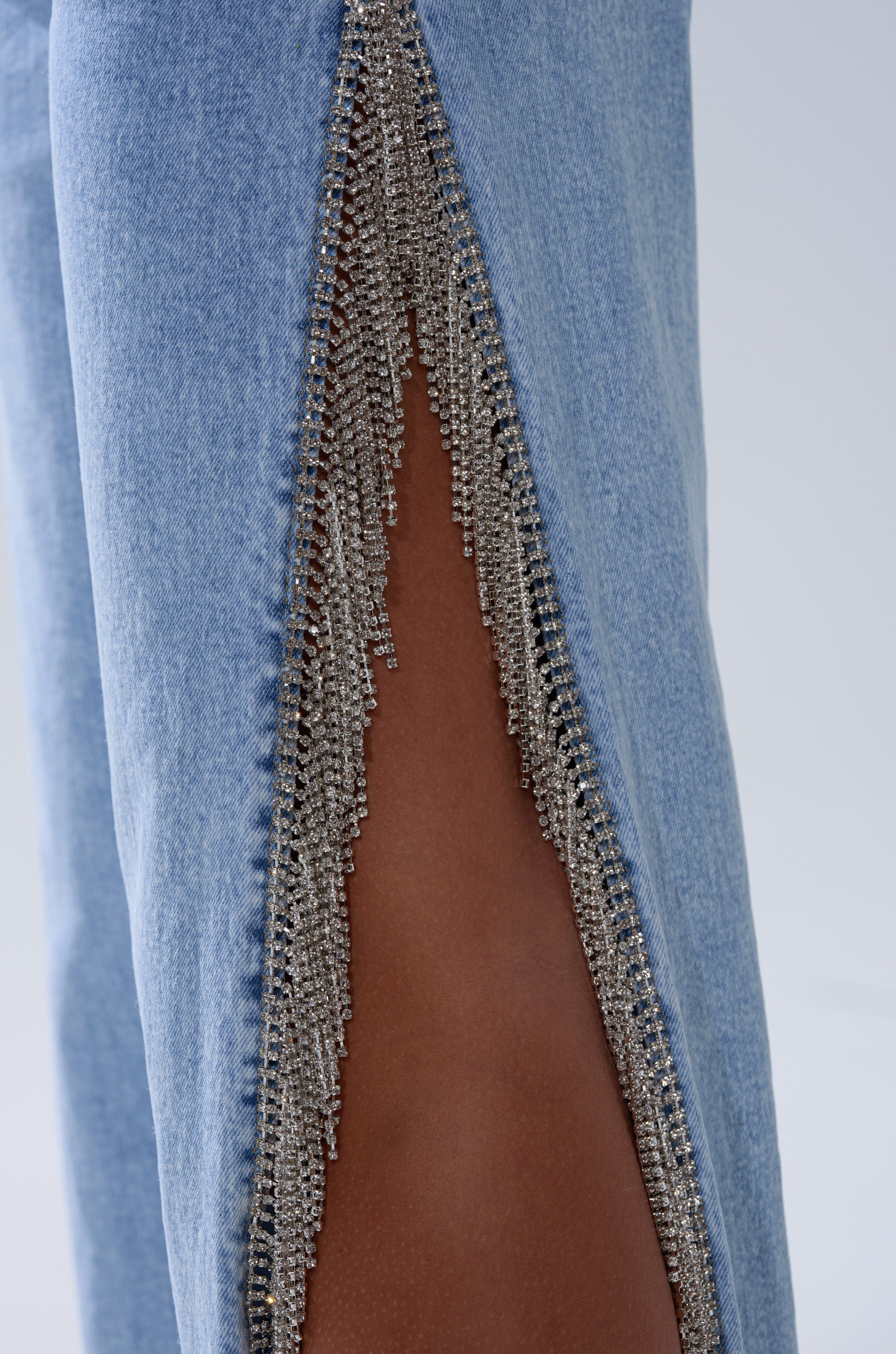 Close-up of ESSENTIAL RELAXED FIT BLING DENIM JEANS in light blue, featuring a high side slit with sparkling rhinestone trim that reveals bare skin underneath.