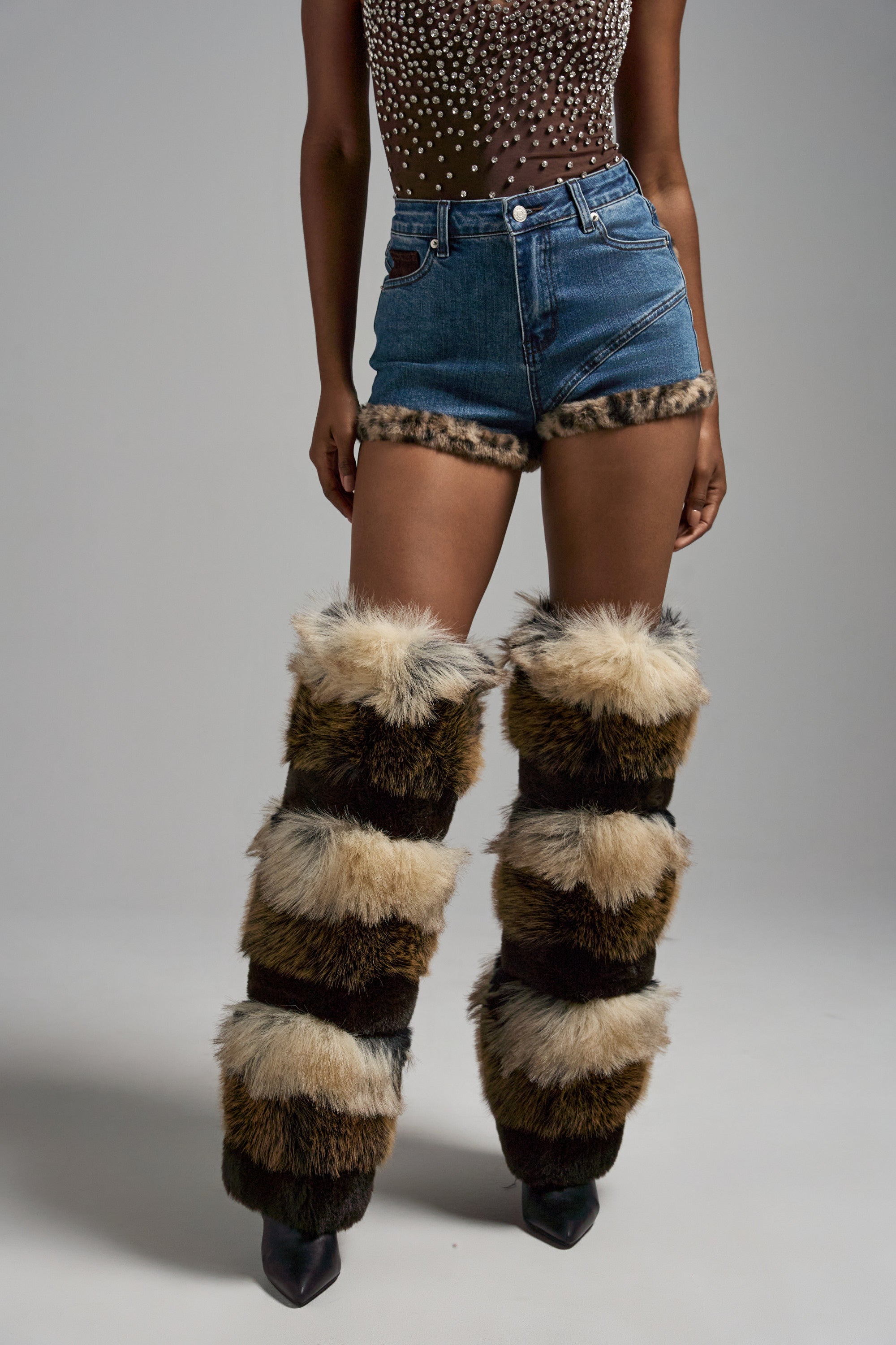 A person wears the LOVE ME MORE DENIM SHORT with leopard-print faux fur trim, a sheer rhinestone top, striped furry leg warmers, and black pointed-toe shoes, standing against a plain gray background. 1