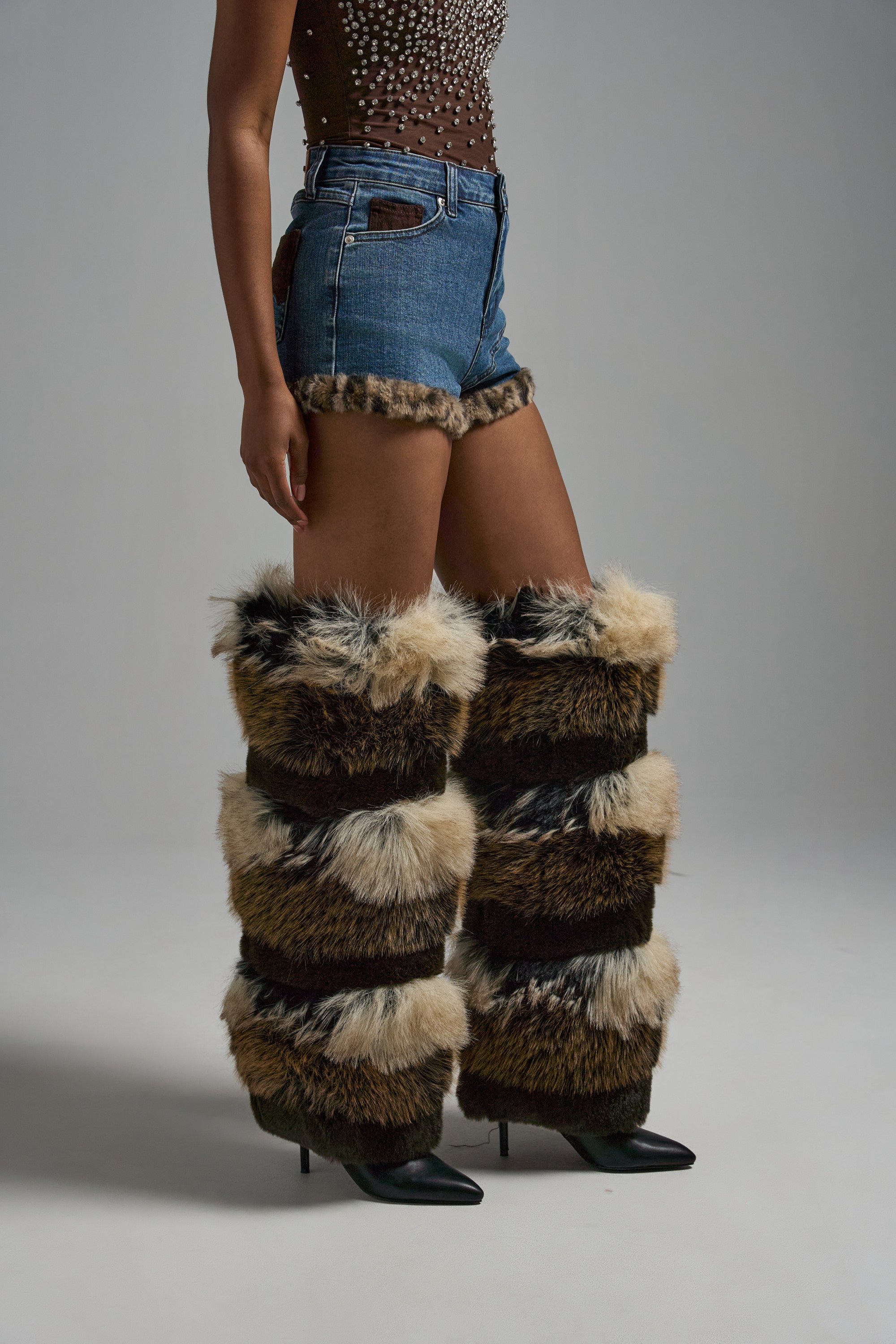 A person wears the LOVE ME MORE DENIM SHORT with leopard-print trim, a brown studded sleeveless top, and tall furry leg warmers over black pointed-toe high-heeled boots, standing against a gray background. 3