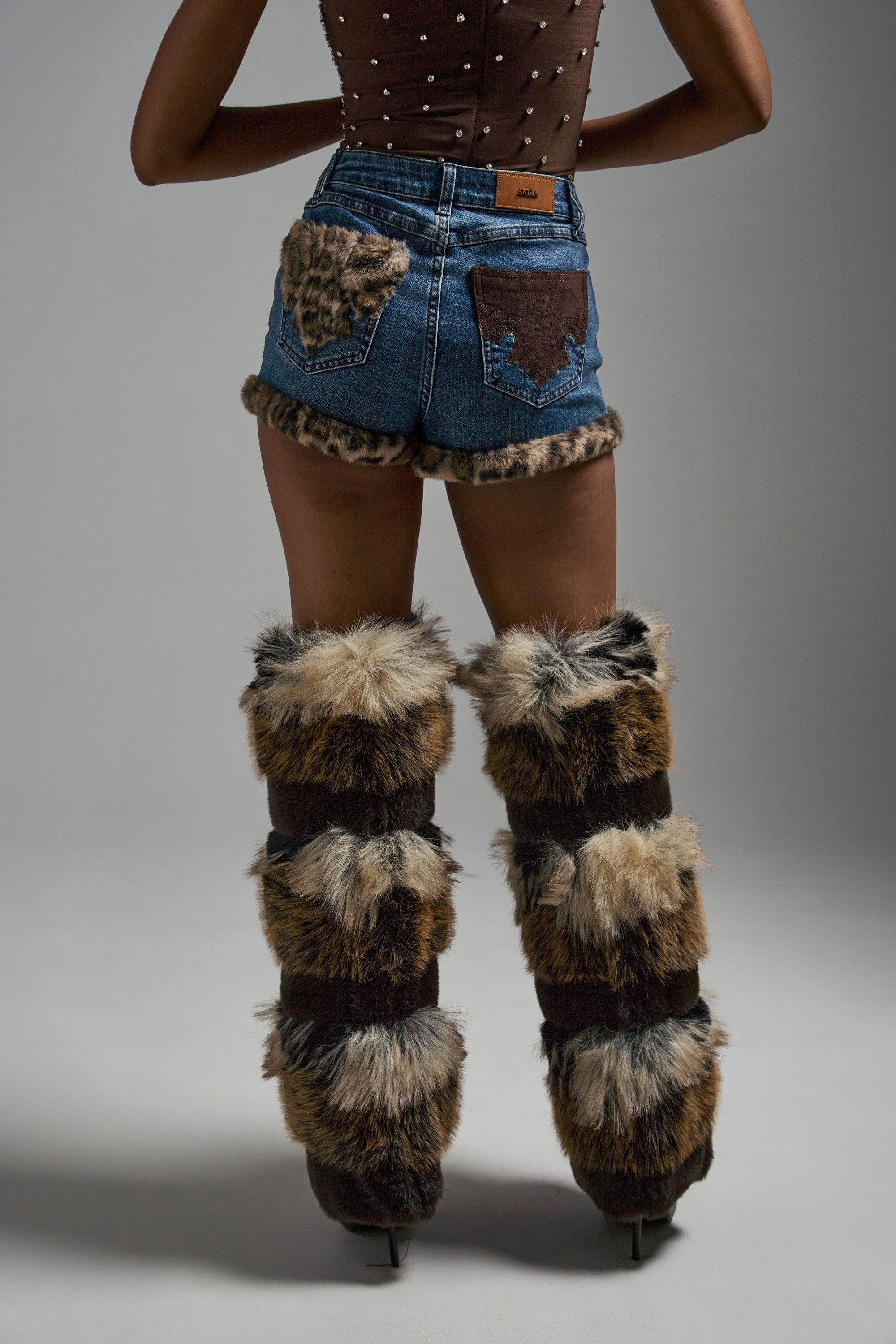 A person faces away, wearing the LOVE ME MORE DENIM SHORT with leopard-print pocket trim, a brown sleeveless top, and tall brown furry thigh-high leg warmers. The backdrop is plain and gray. 0