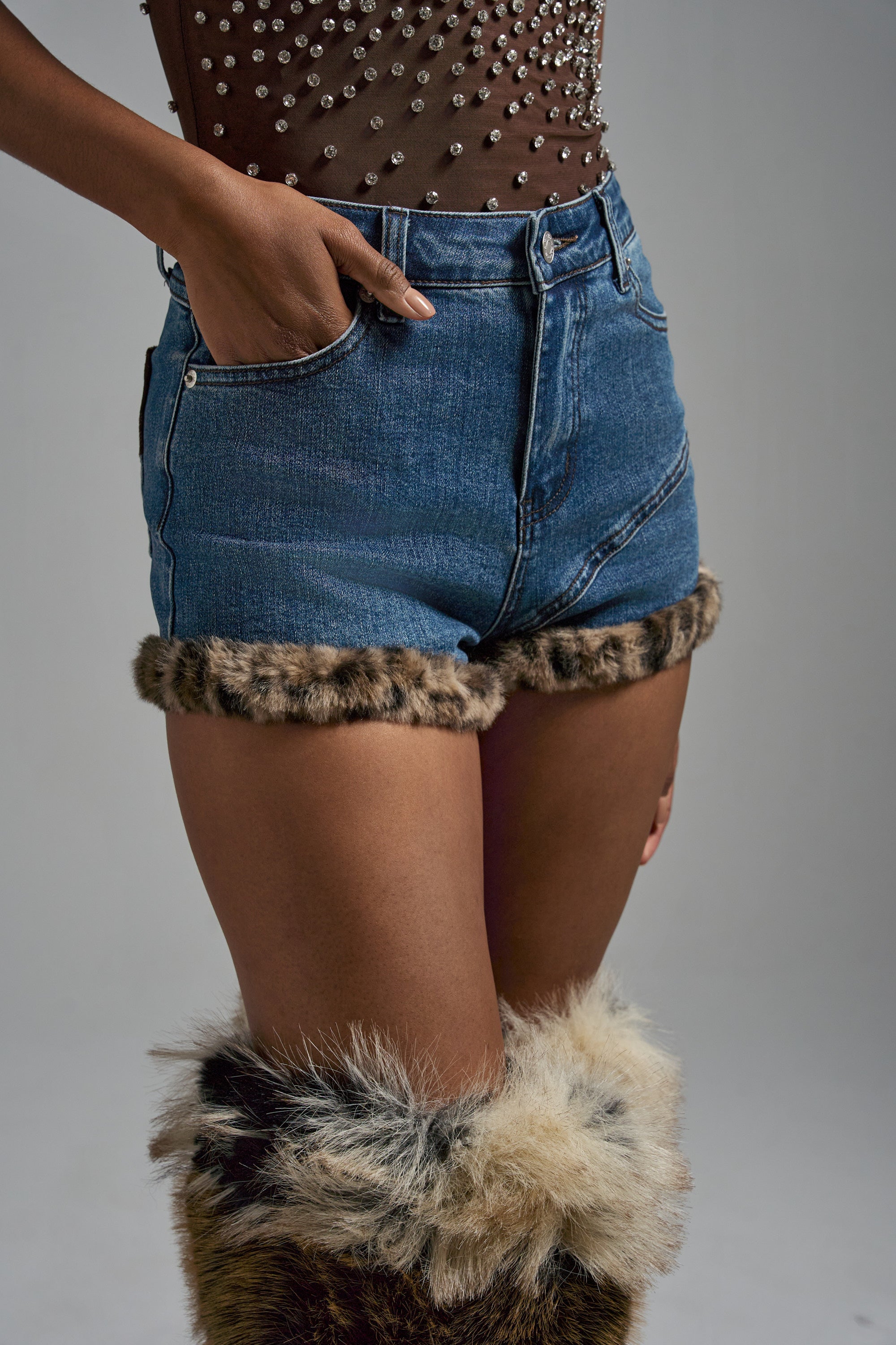 A person wears the LOVE ME MORE DENIM SHORT with leopard-print hem and matching faux fur leg warmers. The sheer brown top is rhinestone-embellished, one hand is in a pocket, and the background is plain gray. 6