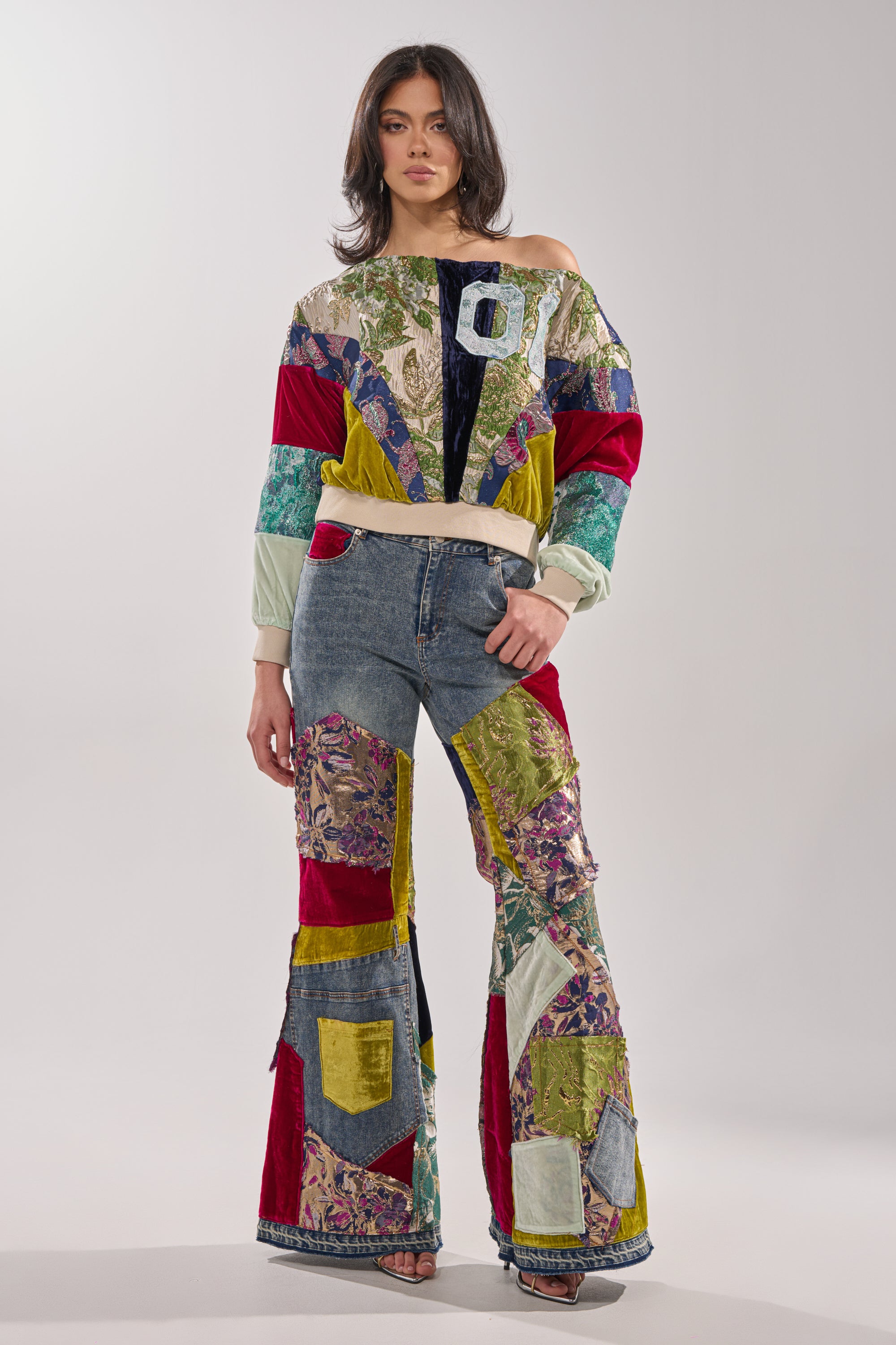 A woman wears the MIXED EMOTIONS BROCADE PATCHWORK FLARE DENIM—high-rise flared jeans and a matching patchwork jacket in assorted colors, velvet, and floral fabrics—posed against a plain light gray background.