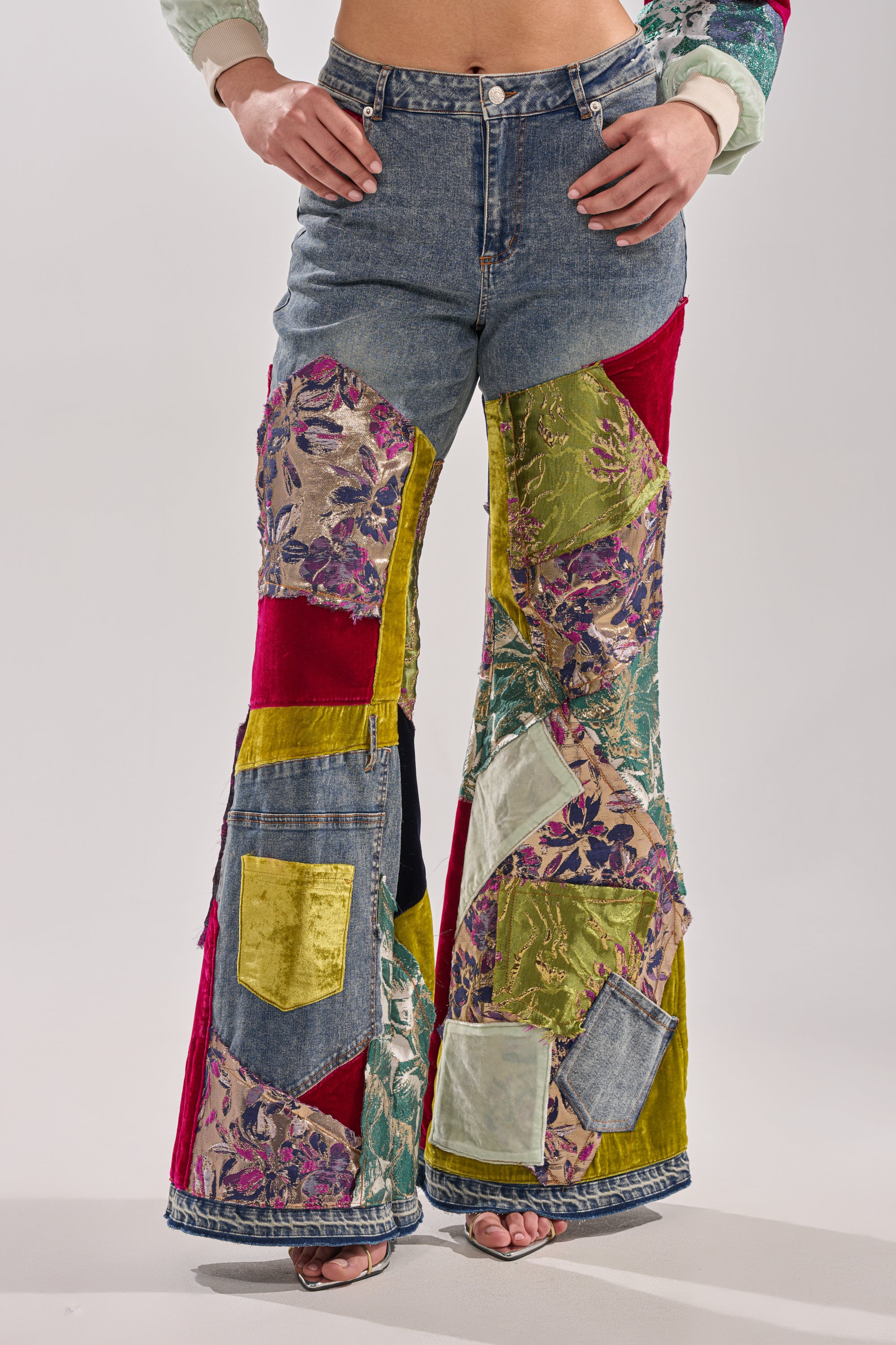 A person wearing the MIXED EMOTIONS BROCADE PATCHWORK FLARE DENIM, featuring high-rise waist and colorful velvet, floral, and metallic brocade patches, stands confidently with hands on hips and feet apart.