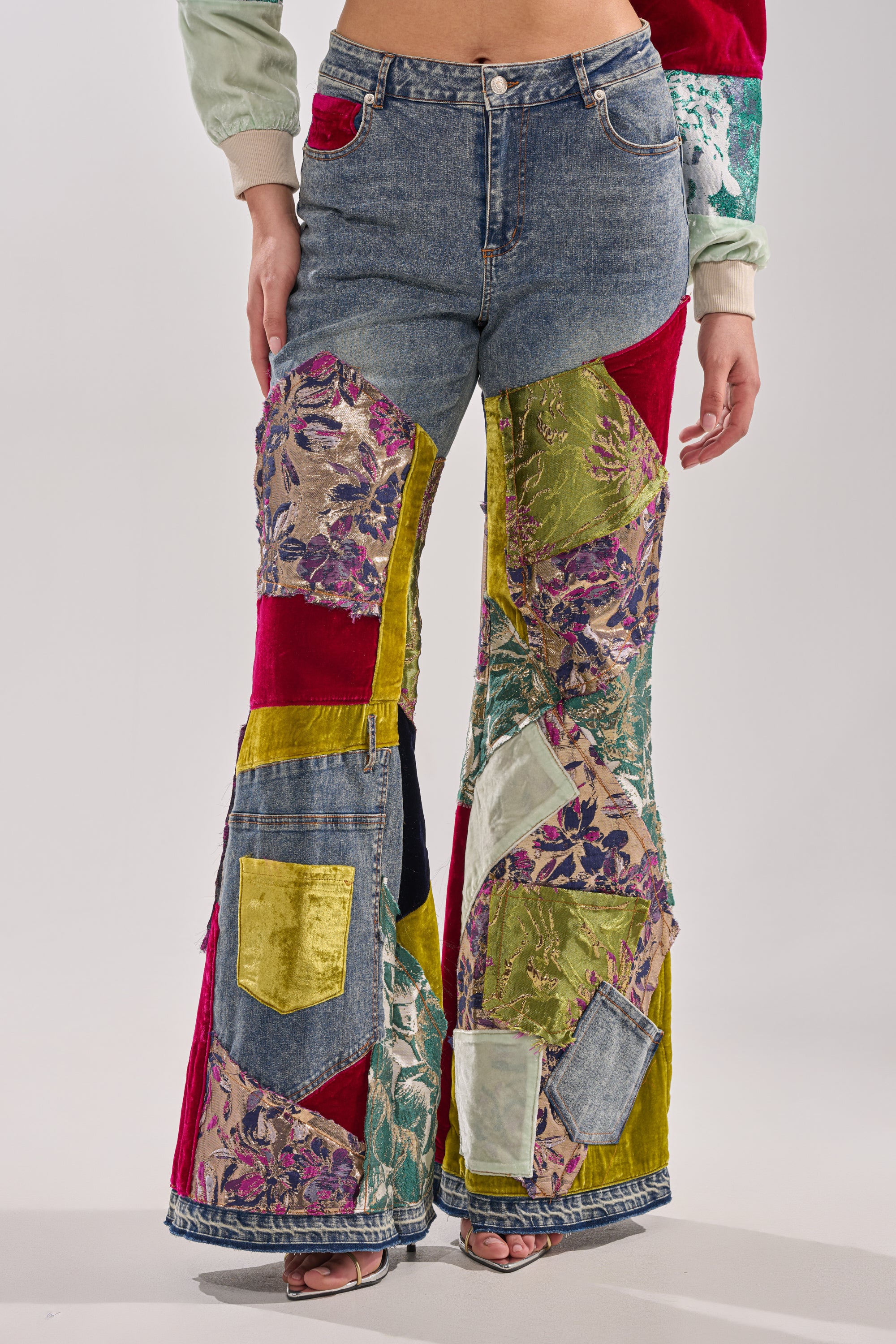 A person wears the MIXED EMOTIONS BROCADE PATCHWORK FLARE DENIM, featuring a high-rise waist and colorful brocade patches, paired with open-toe heels. The straight-to-flared legs are shown from waist to ankles in the cropped image.