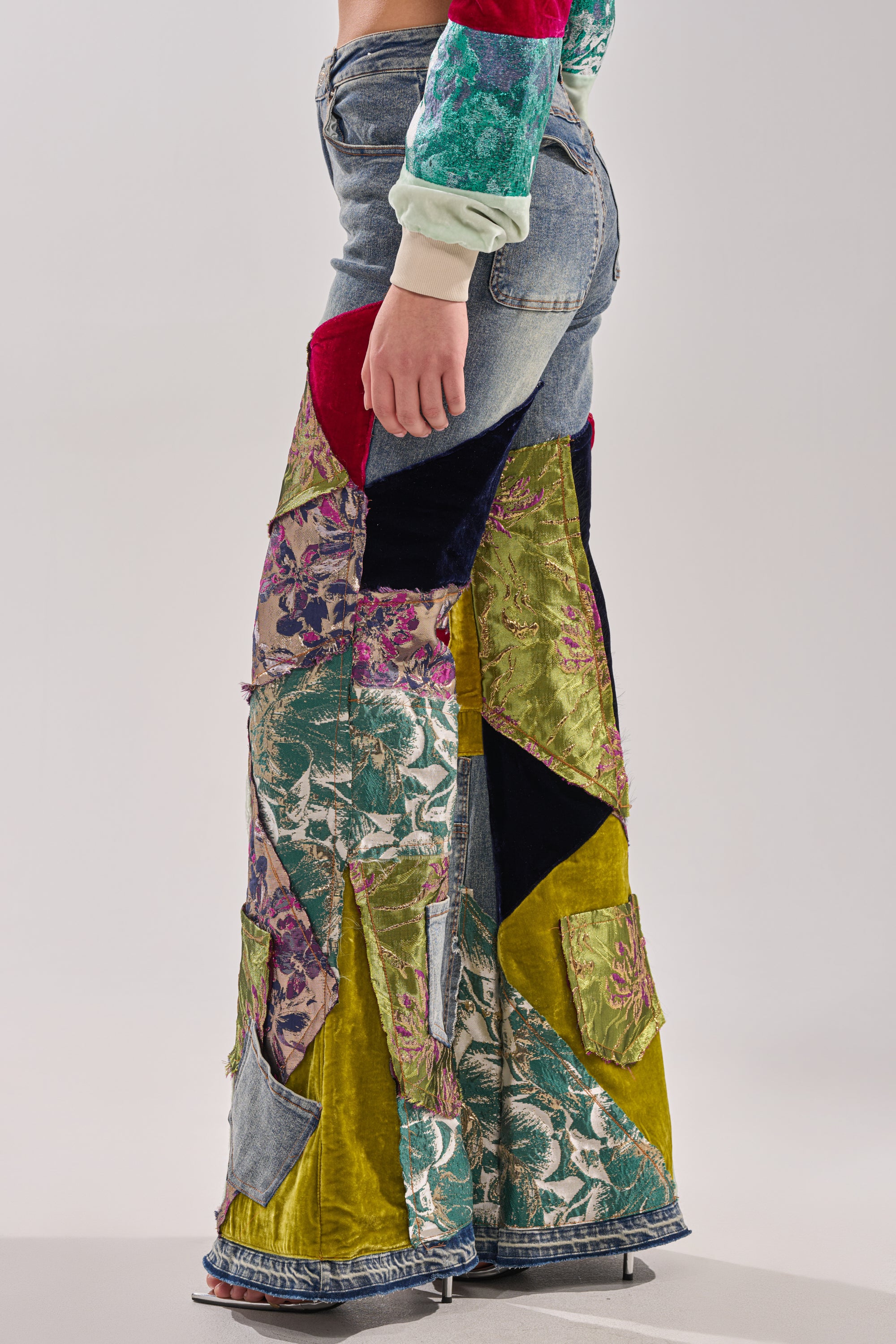 A person wearing the MIXED EMOTIONS BROCADE PATCHWORK FLARE DENIM—high-rise, wide-leg jeans in patchwork denim, velvet, and floral brocade—stands sideways against a plain light background.