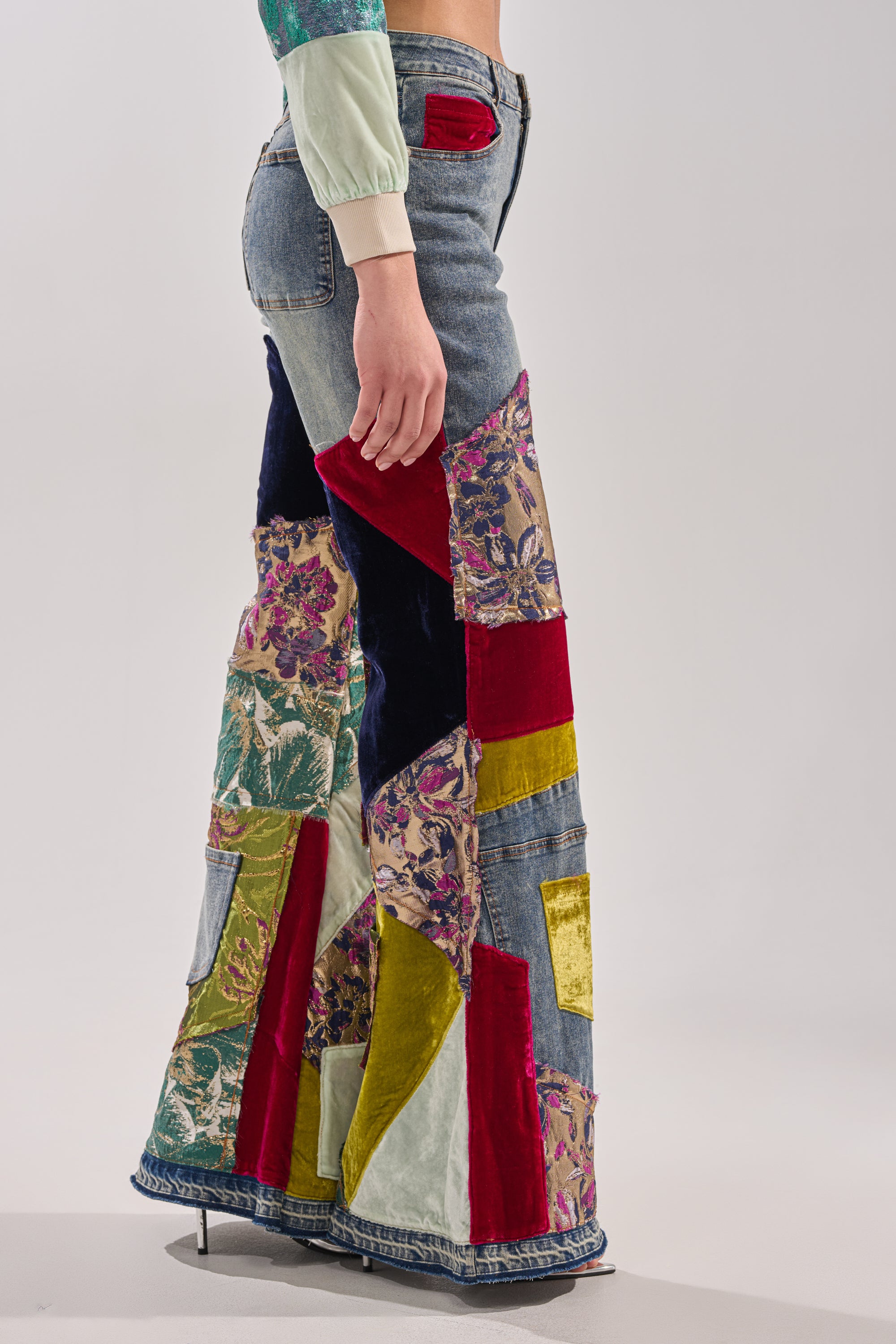 A person in profile wears the MIXED EMOTIONS BROCADE PATCHWORK FLARE DENIM, featuring high-rise waist and colorful floral, velvet, and denim patches, set against a neutral background.