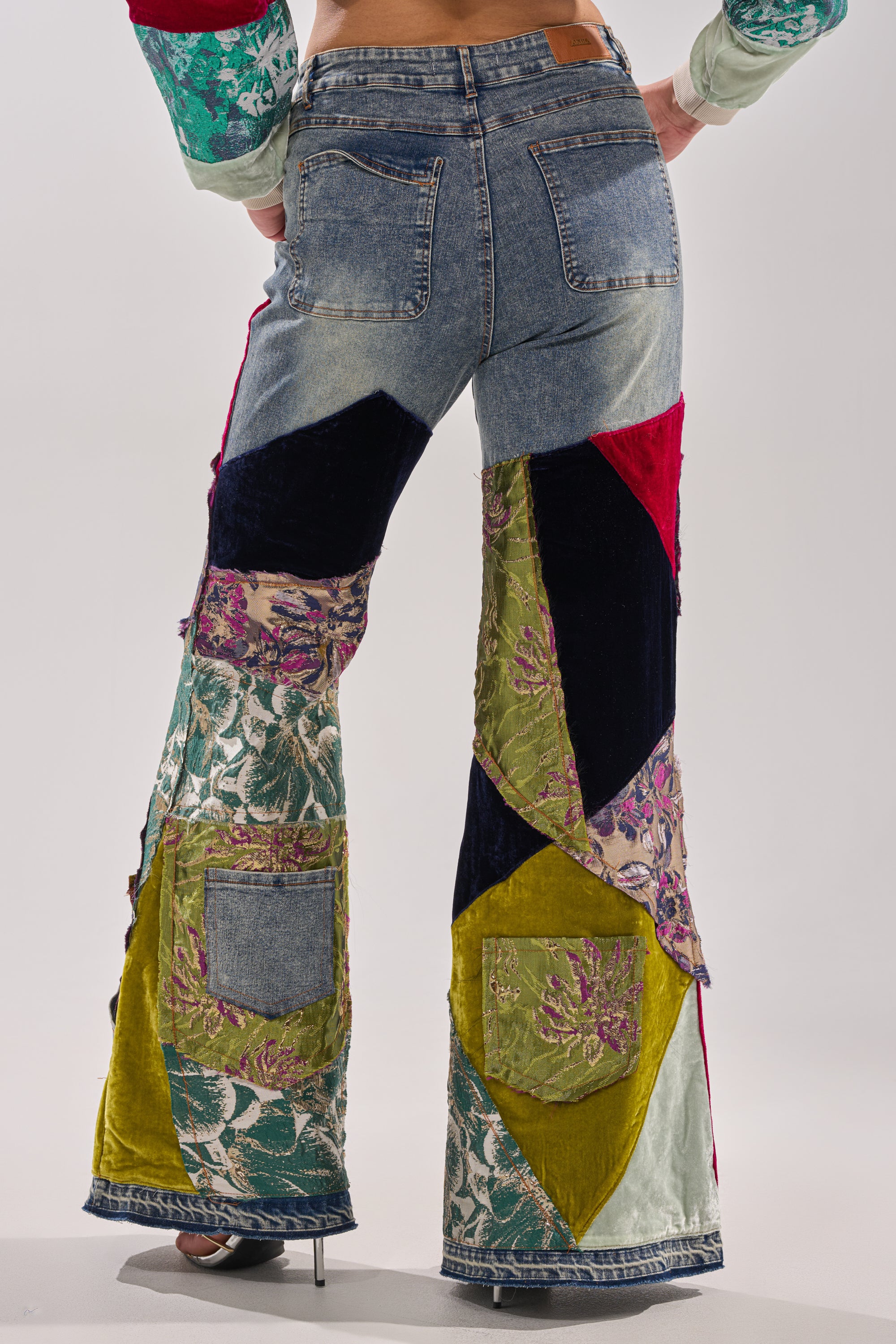 A person wears the MIXED EMOTIONS BROCADE PATCHWORK FLARE DENIM, featuring a high-rise fit and flared legs with blue denim, green velvet, red, navy, and floral fabrics. Photo shows the back view from waist to feet.