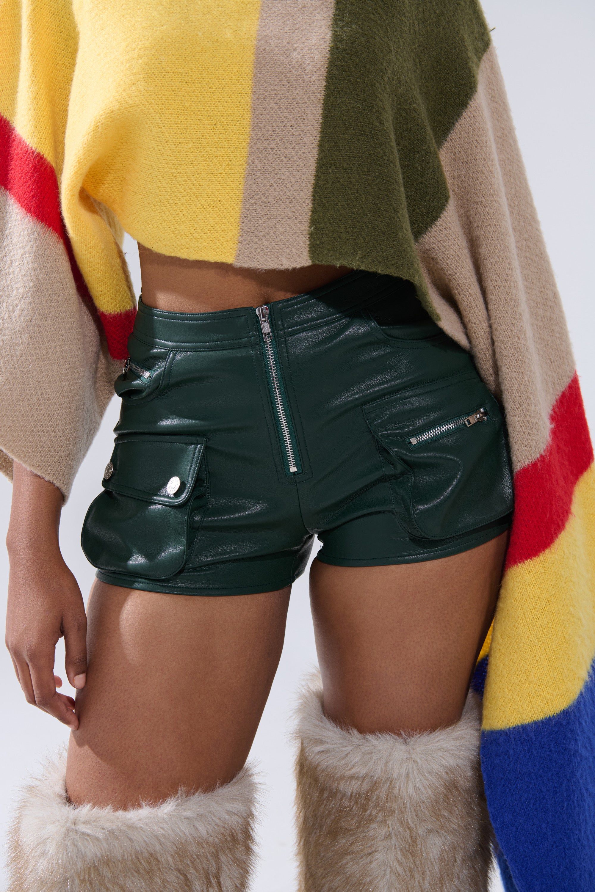 Someone is wearing FAUX LEATHER MINI CARGO SHORTS in dark green high-waisted style, a vibrant striped poncho with yellow, green, red, and blue, and tan fur-trimmed knee-high boots. Their face isn't visible. 0