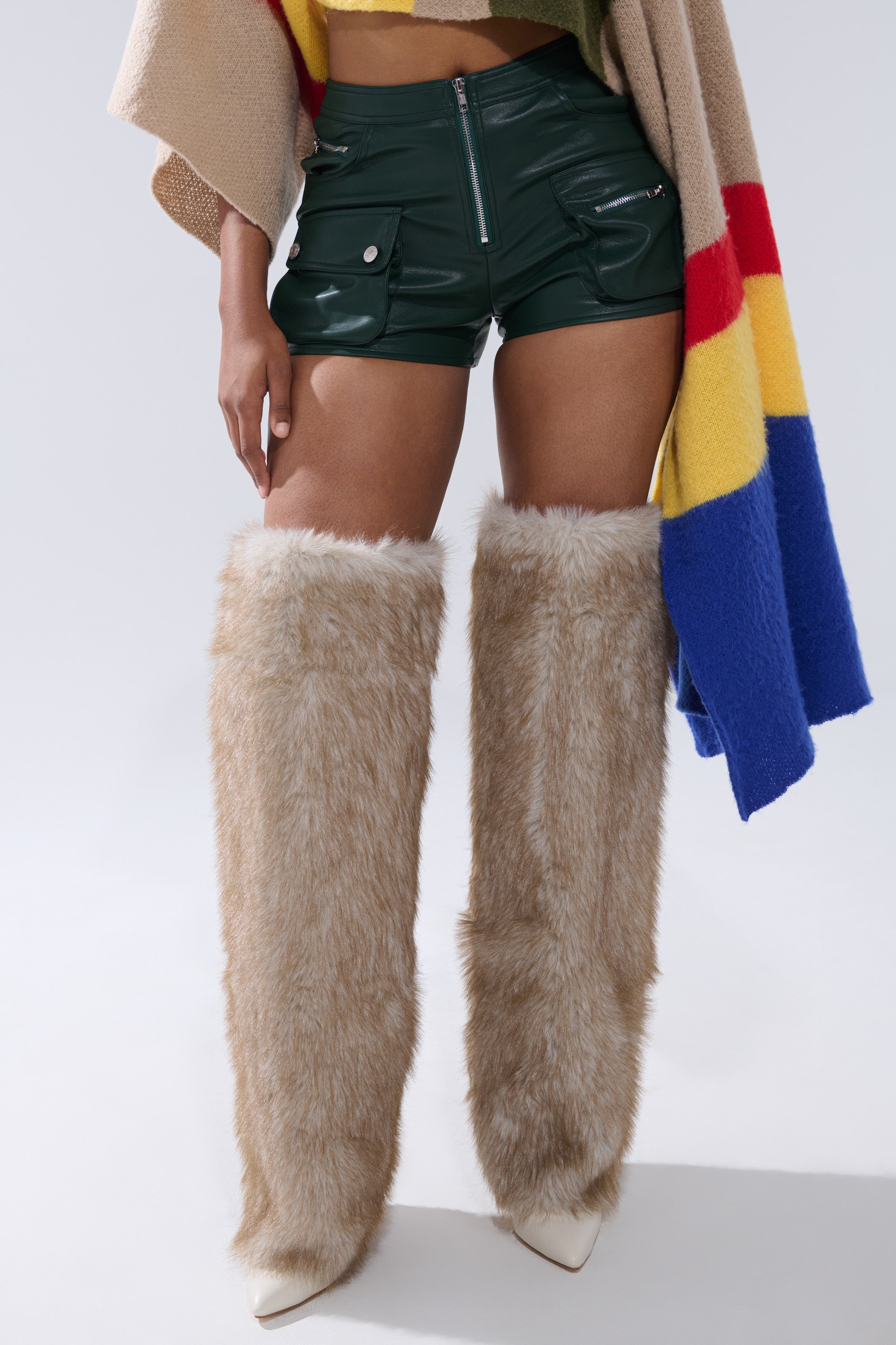 A person wearing shiny dark green FAUX LEATHER MINI CARGO SHORTS, a tan and multicolored poncho, and tall, tan faux fur boots that cover the thighs, paired with pointed white heels. 1