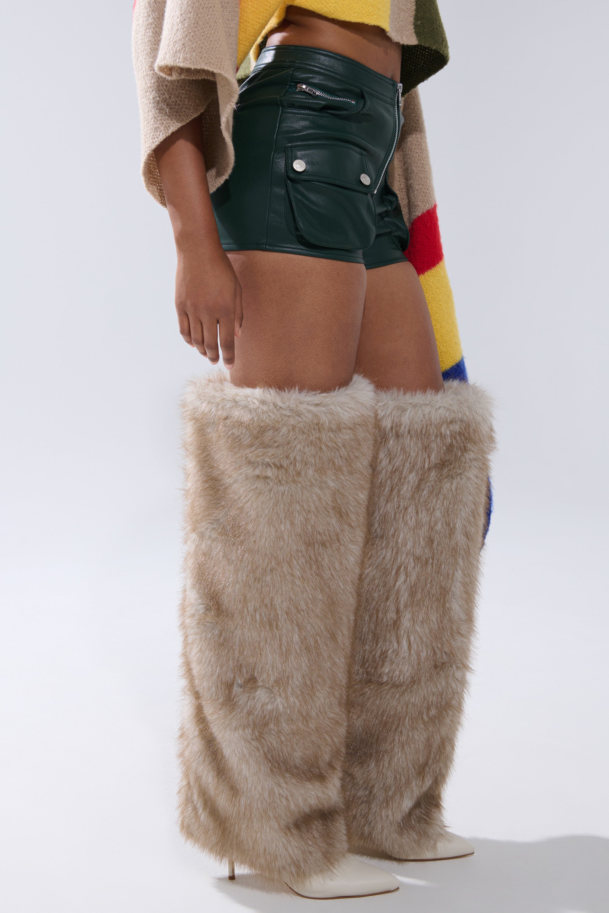 A person wears dark green FAUX LEATHER MINI CARGO SHORTS, a colorful striped sweater, and tall beige faux fur boots with white pointed heels, standing against a plain light background. 2