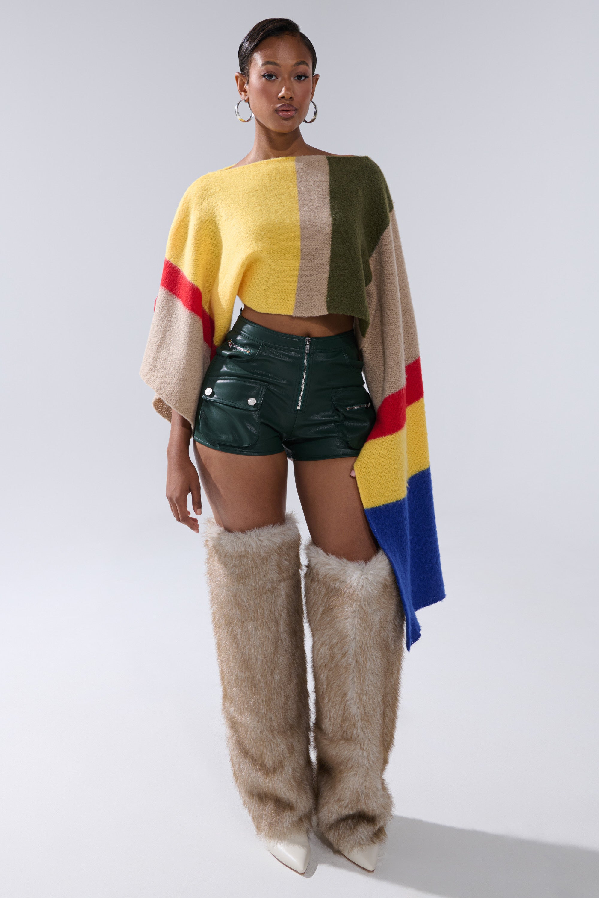 A woman wears FAUX LEATHER MINI CARGO SHORTS in dark green, paired with a color-block poncho, large hoop earrings, and knee-high light brown furry boots. She stands against a plain, light grey background. 4