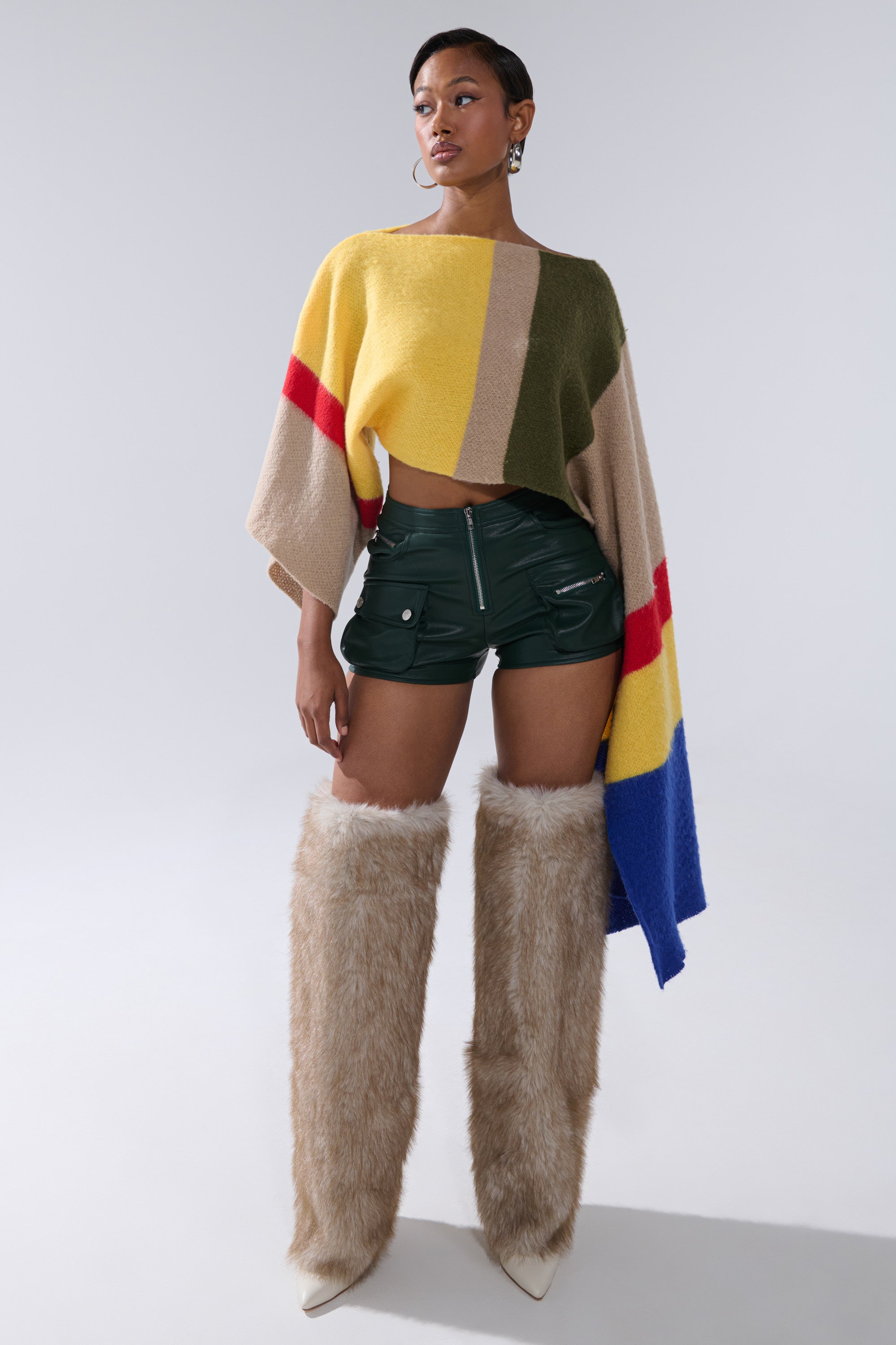 A woman stands confidently in colorful, geometric-patterned knitwear, FAUX LEATHER MINI CARGO SHORTS, and tall beige furry boots against a plain gray background. 5