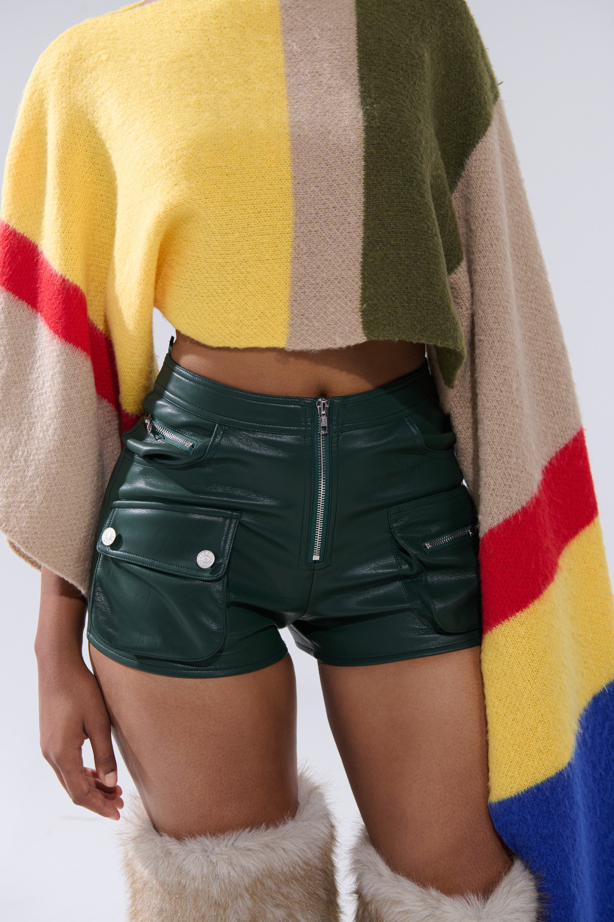 A person models FAUX LEATHER MINI CARGO SHORTS with front zippers and pockets, paired with a color-blocked poncho and furry knee-high boots. The photo focuses on the outfit from waist to mid-thigh. 6
