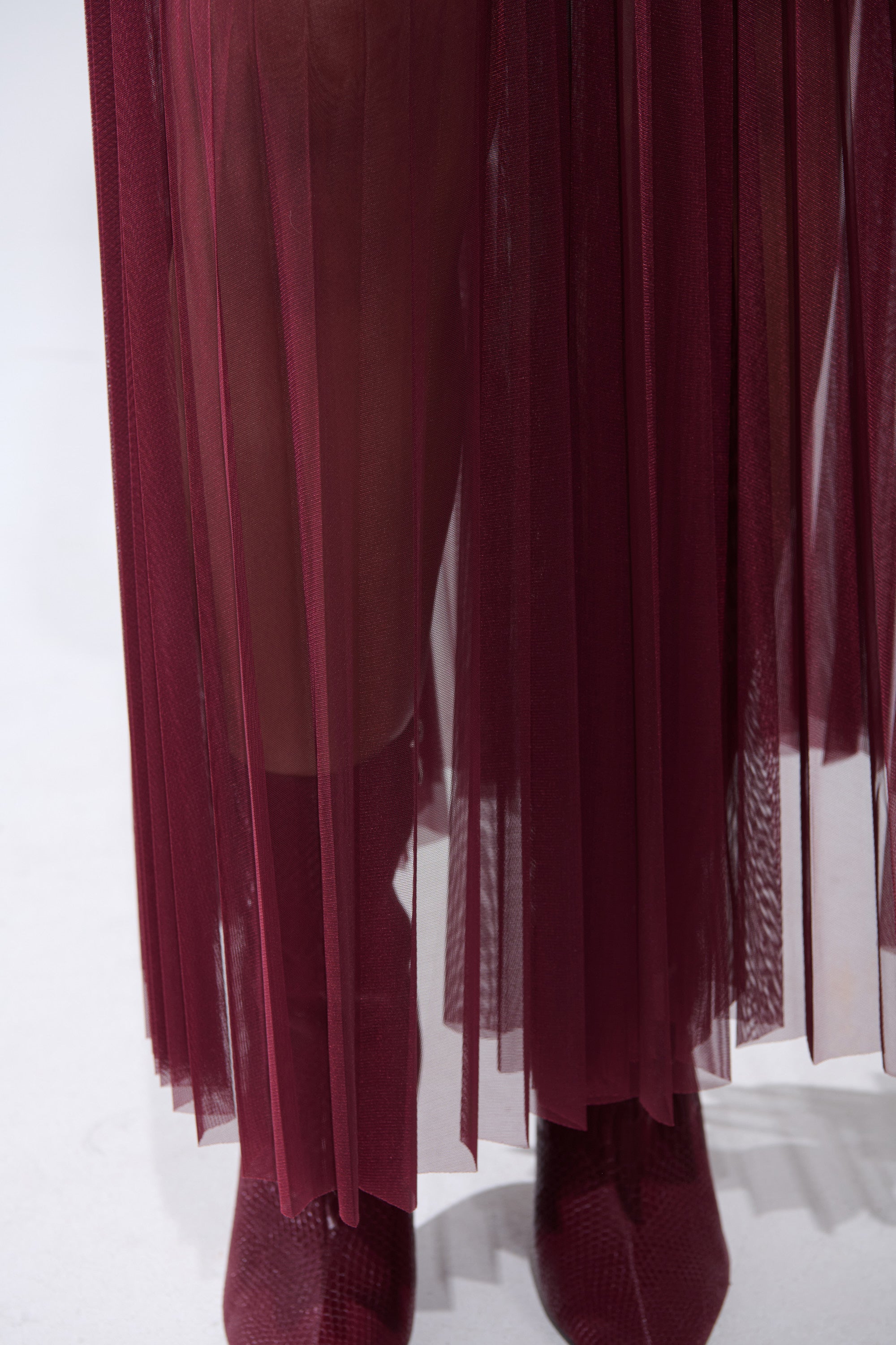 A close-up of a person wearing the ON FIRE MESH MAXI SKIRT in sheer burgundy over their legs, paired with matching textured burgundy ankle boots against a light background. 9