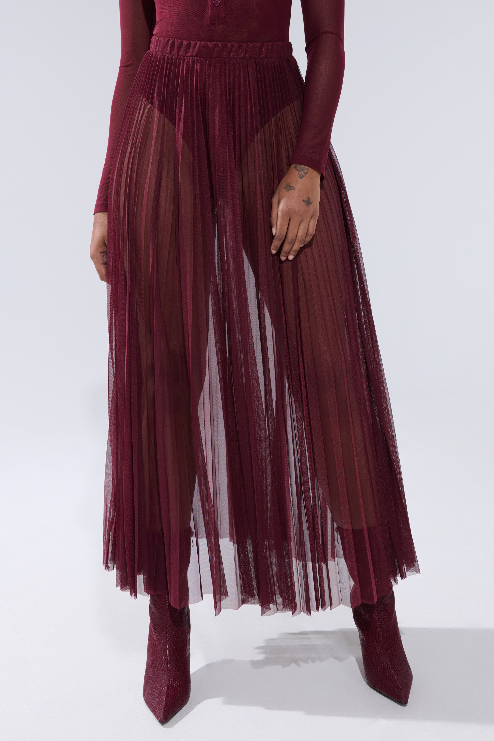 A person wears the ON FIRE MESH MAXI SKIRT with a matching long-sleeve top and pointed boots. Their tattooed hand rests by their side, set against a plain light background, highlighting this elegant mesh skirt ensemble. 2