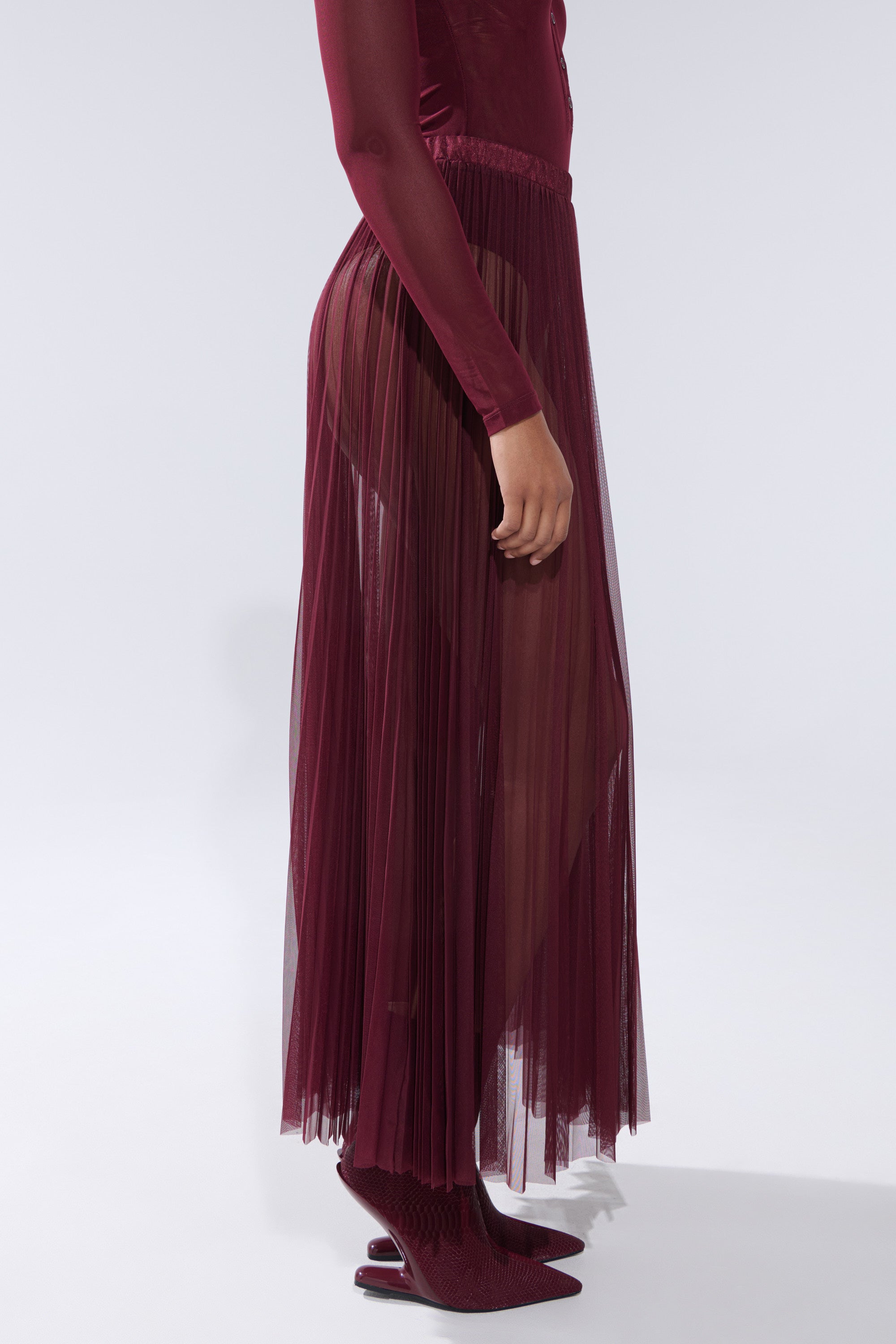A person stands in profile against a plain light background, wearing the ON FIRE MESH MAXI SKIRT in burgundy, paired with a fitted long-sleeved top and matching pointed-toe heels. 3