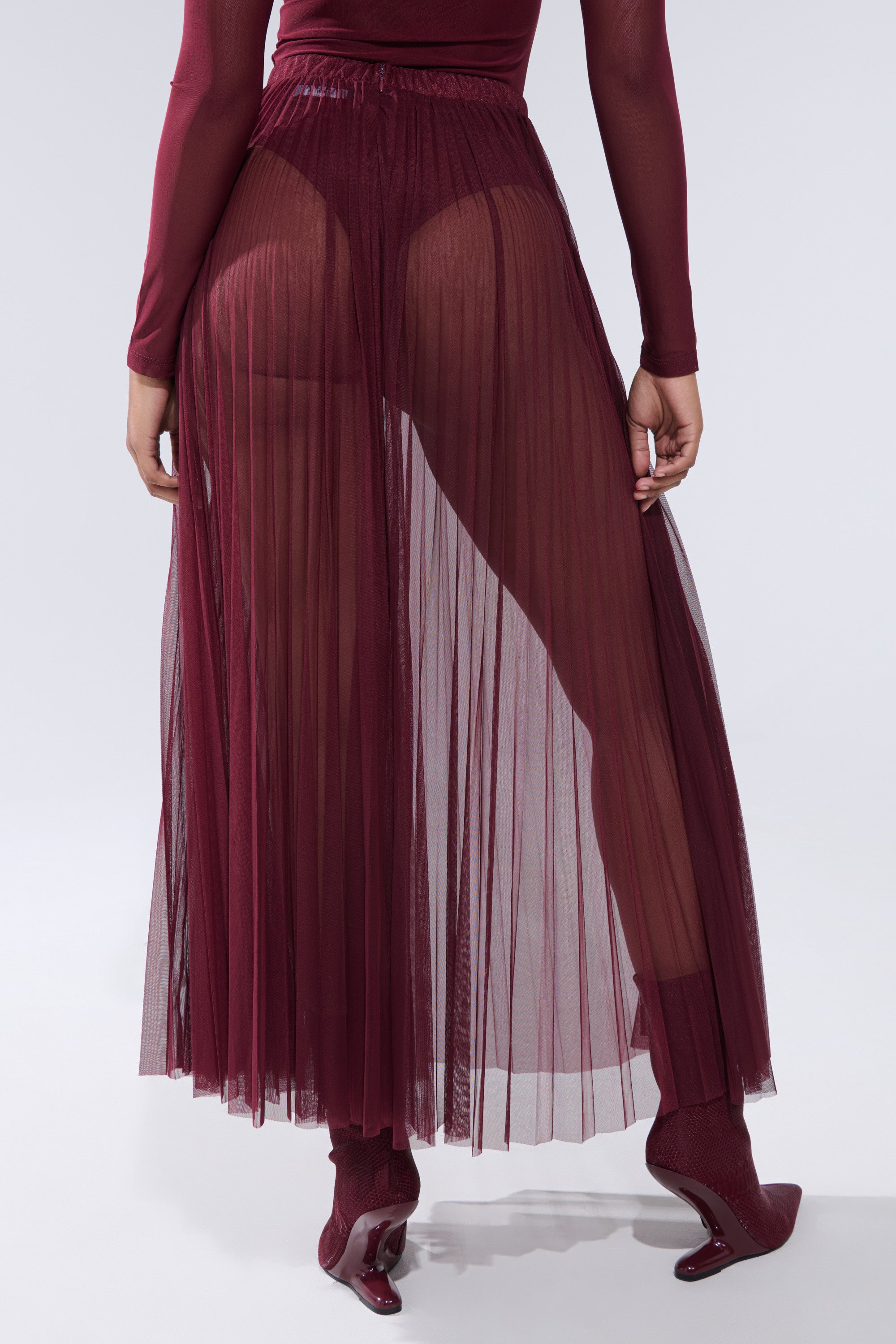 Model wears the ON FIRE MESH MAXI SKIRT, a sheer burgundy mesh maxi skirt layered over matching high-waisted undergarments, paired with a long-sleeve top, socks, and heels for a bold semi-transparent look that highlights the leg silhouette. 4