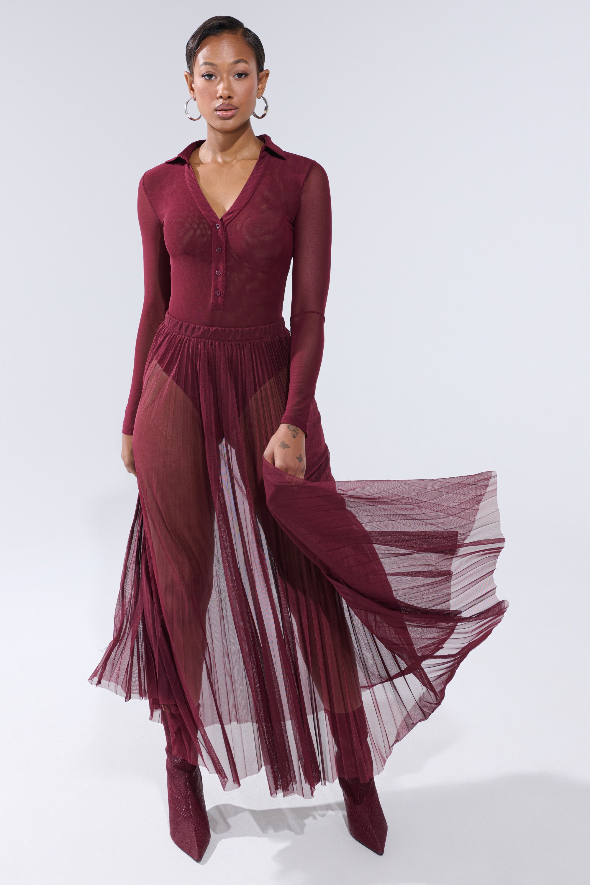 A woman models the ON FIRE MESH MAXI SKIRT—a sheer, pleated burgundy maxi with a collared top and long sleeves—styled with matching boots against a light background. 6