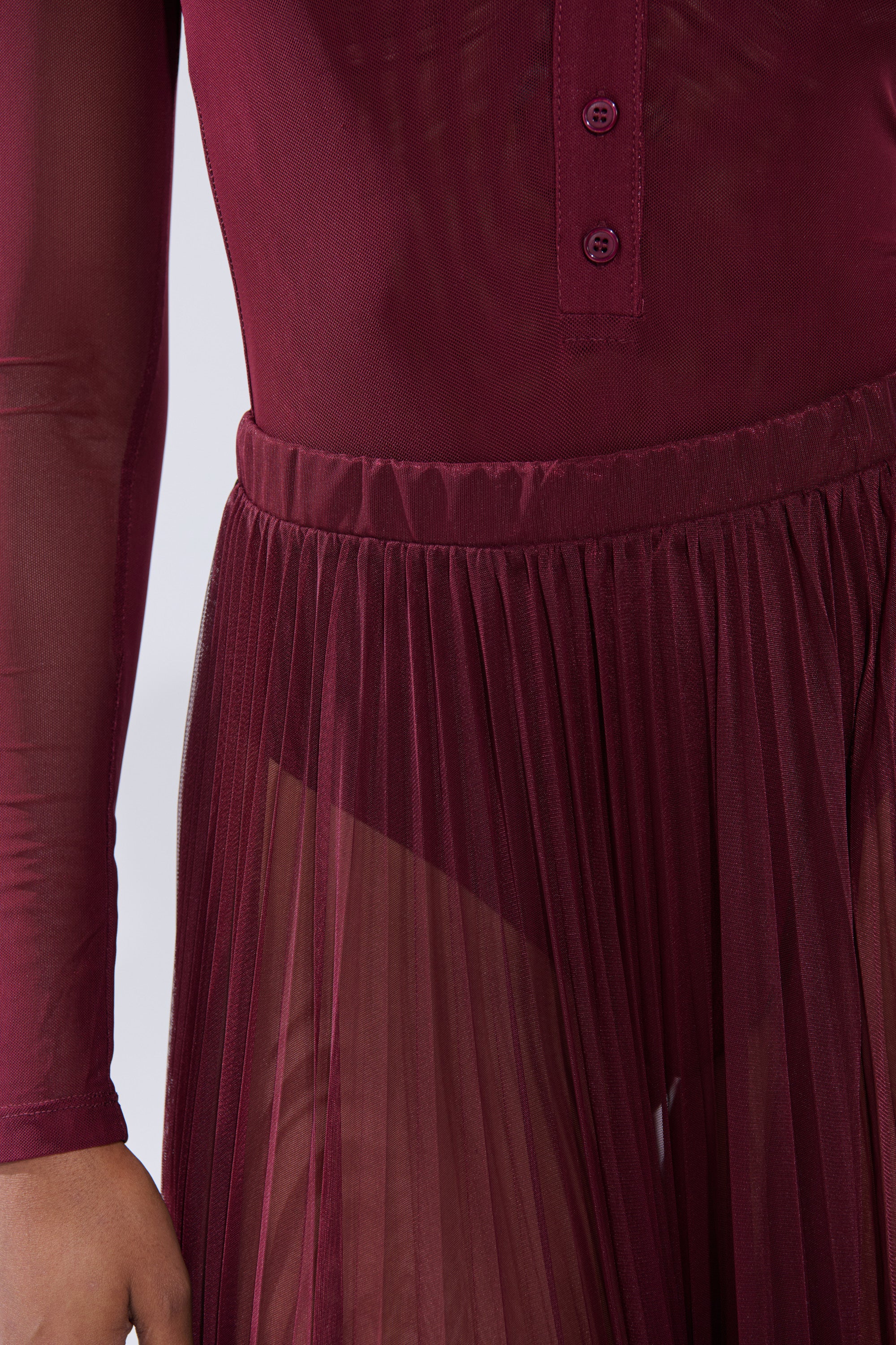 A close-up of a person wearing the ON FIRE MESH MAXI SKIRT, featuring sheer burgundy mesh, long sleeves, a buttoned top, and a pleated maxi silhouette with semi-transparent fabric that subtly reveals undergarments. 8