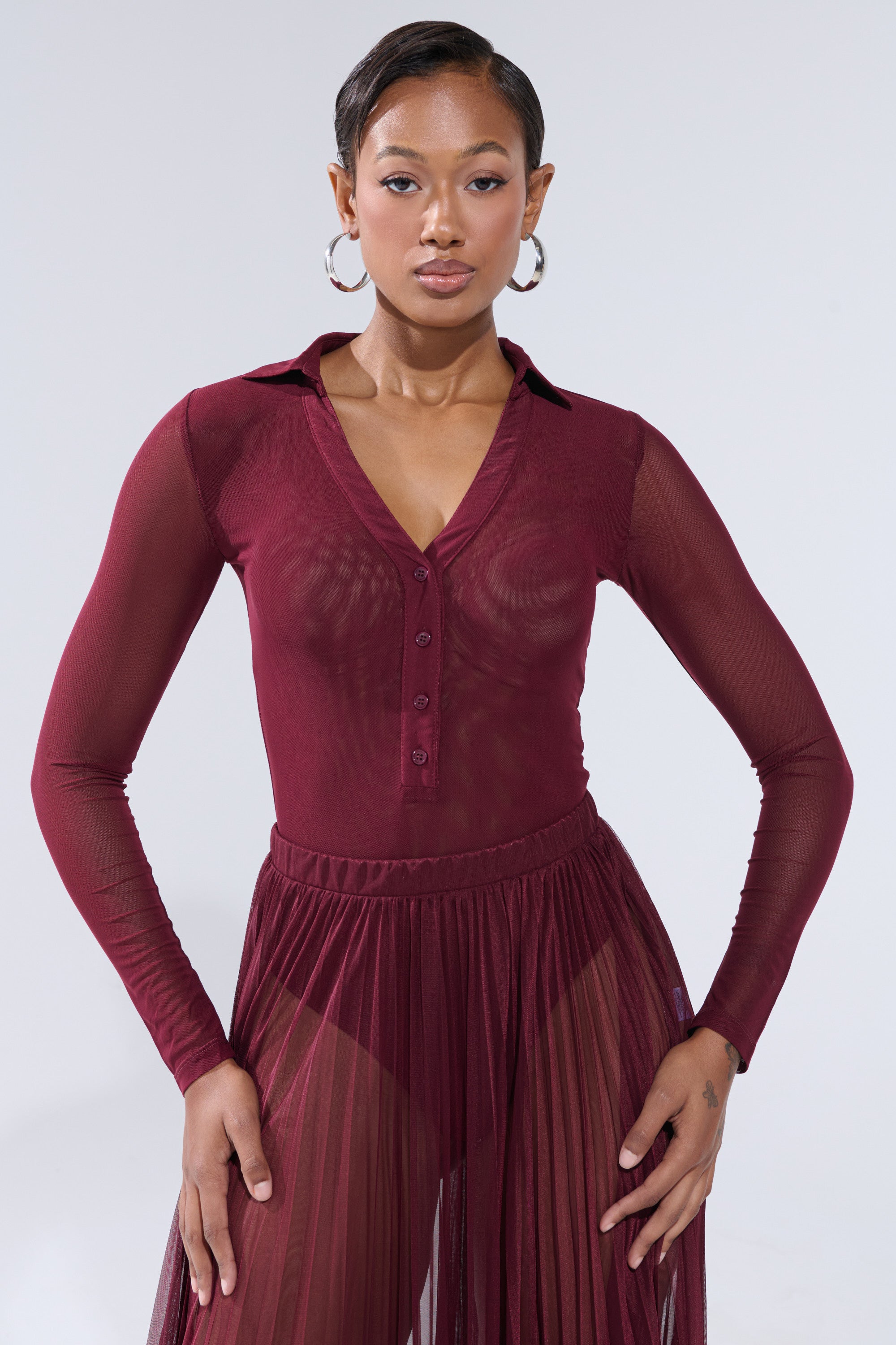A woman stands confidently in the ON FIRE MESH BODYSUIT by AKIRA Label, styled with a sheer burgundy pleated skirt, hoop earrings, and slicked-back hair against a plain background. 0