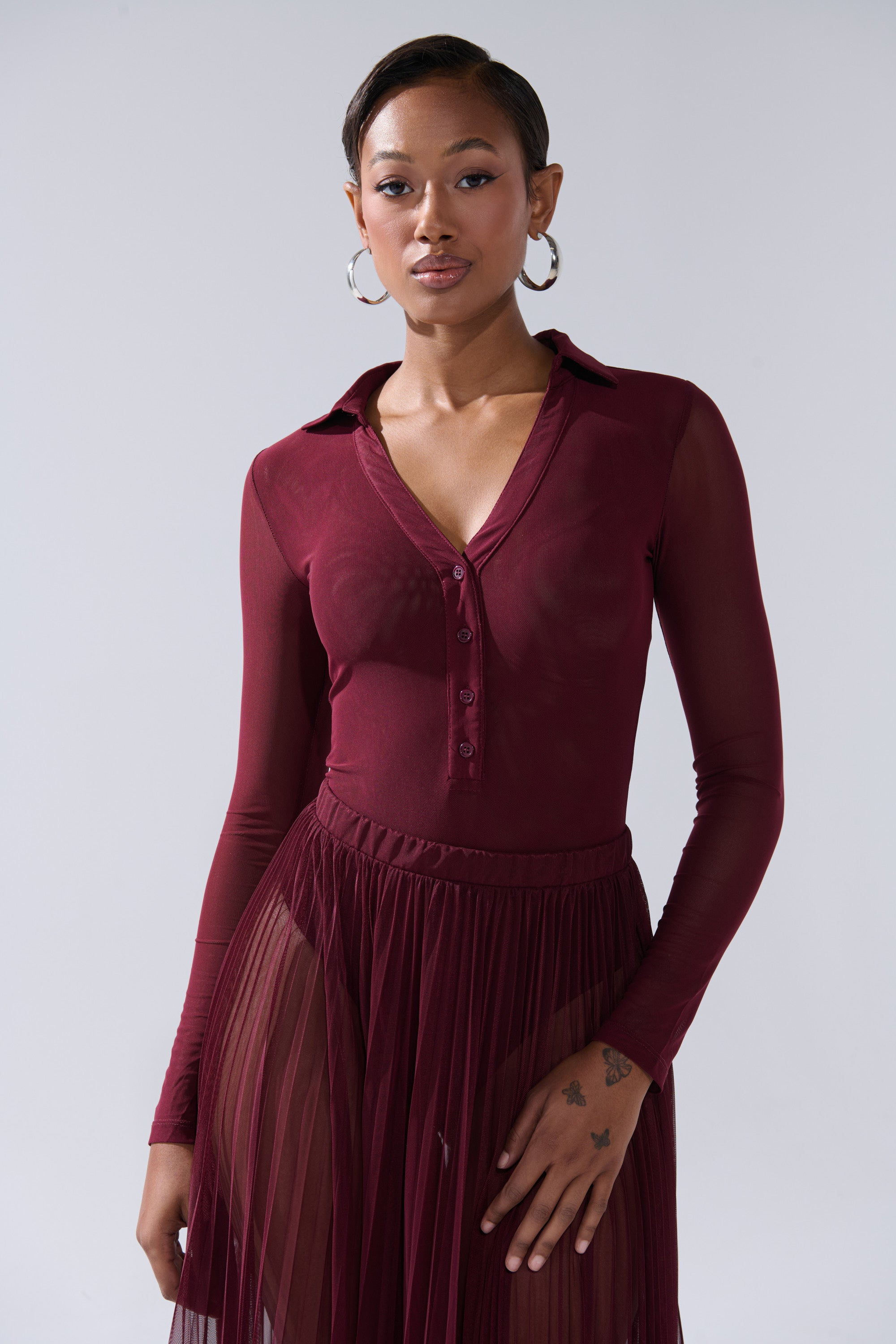 A woman with short hair, hoop earrings, and a tattoo on her left hand poses against a light background in the ON FIRE MESH BODYSUIT—a sheer burgundy, long-sleeve, curve-hugging top with buttons—paired with a matching pleated skirt. 1
