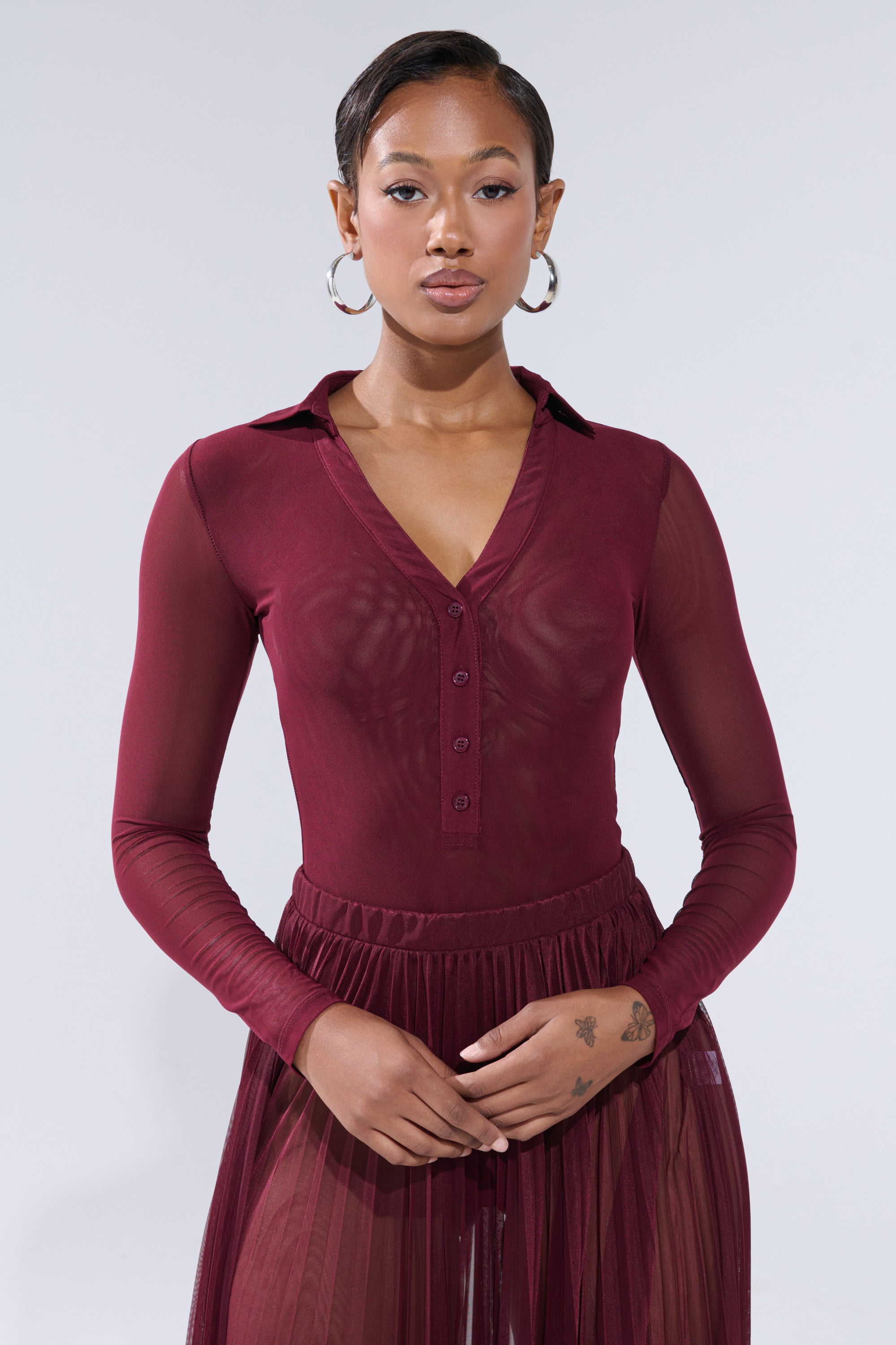 A woman models the ON FIRE MESH BODYSUIT—a sheer, long-sleeved mesh style with a collar and buttons—standing confidently against a plain background with her hands folded in front. 2