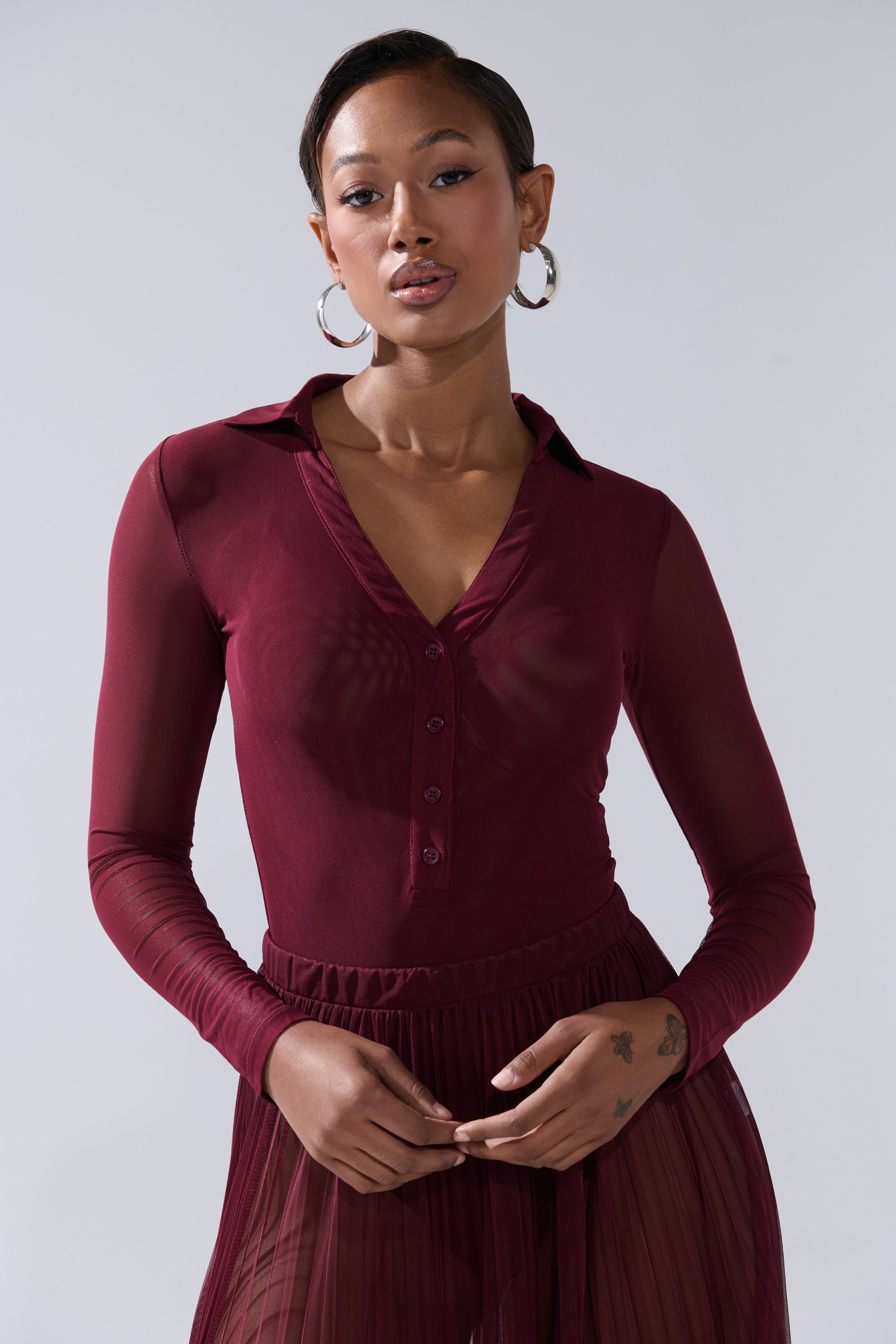 A woman with short hair, hoop earrings, and a tattoo on her left wrist stands against a light background wearing the ON FIRE MESH BODYSUIT, a sheer long-sleeve bodysuit featuring a plunging V-neckline and curve-hugging fit. 3