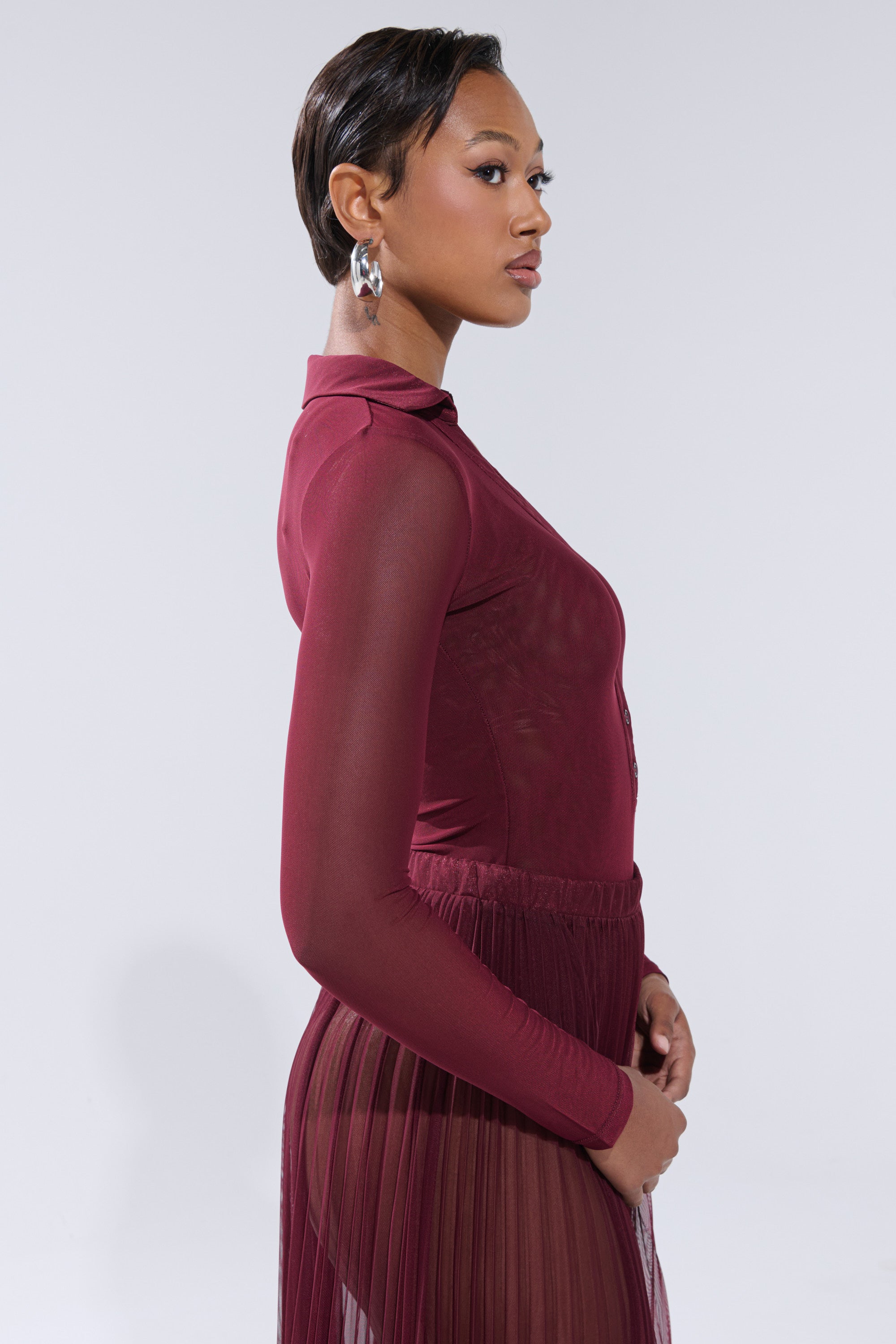 A woman with short hair wears the ON FIRE MESH BODYSUIT in burgundy and a matching pleated skirt, posing in profile against a plain light gray background with a neutral expression. 4