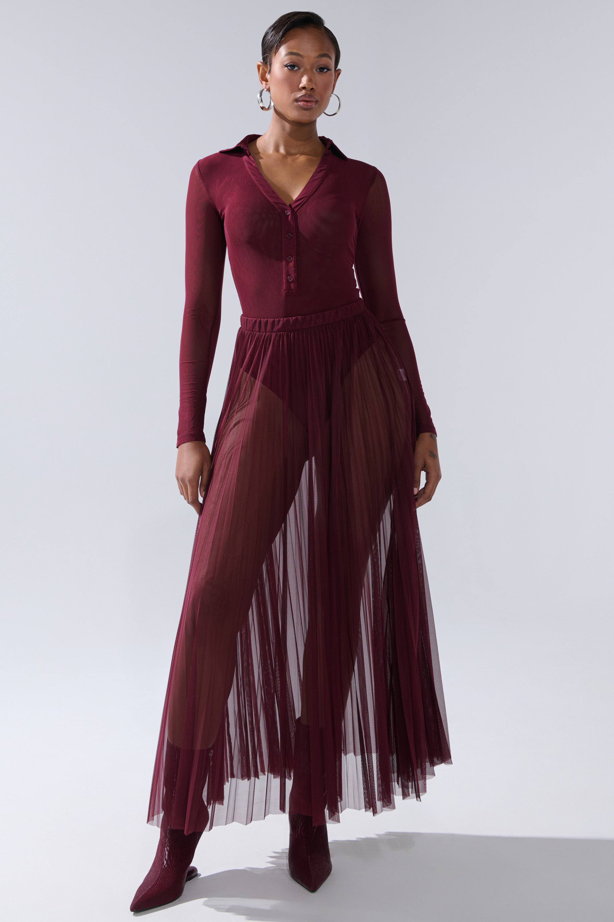 A woman stands confidently against a plain light background wearing the ON FIRE MESH BODYSUIT in burgundy with a curve-hugging fit and pleated skirt, paired with matching pointed boots and hoop earrings. 6
