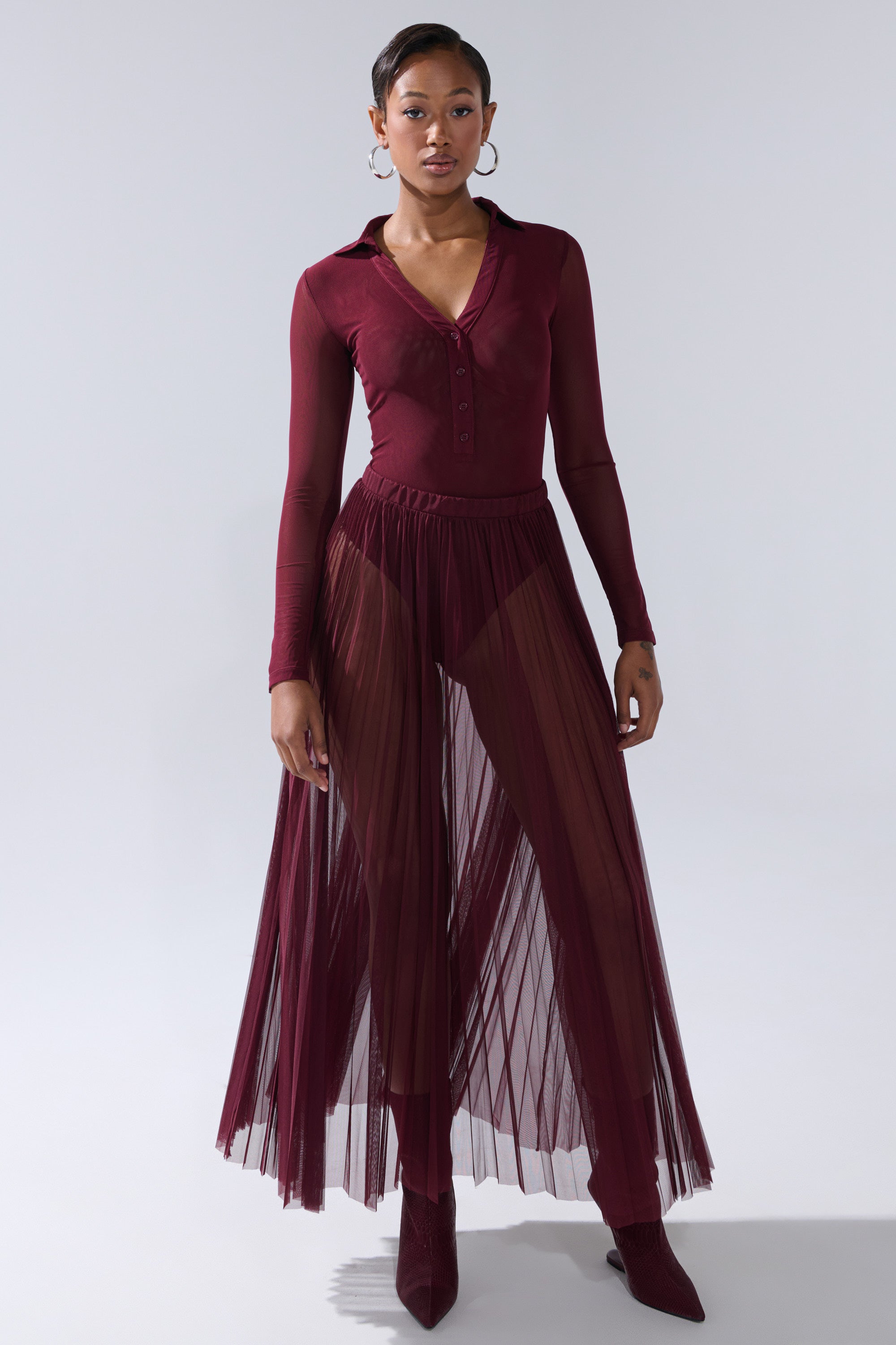 A woman exudes confidence in the ON FIRE MESH BODYSUIT, paired with a matching pleated tulle maxi skirt, opaque shorts, pointed boots, and hoop earrings, posing with a neutral expression against a plain background. 8