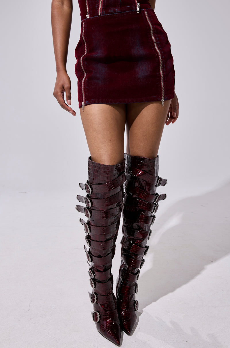 A person wears the FOLD OVER ZIP MINI SKIRT in dark red, styled with matching knee-high, crocodile-patterned boots with multiple buckles and pointed toes. The photo features a plain, light background. 4