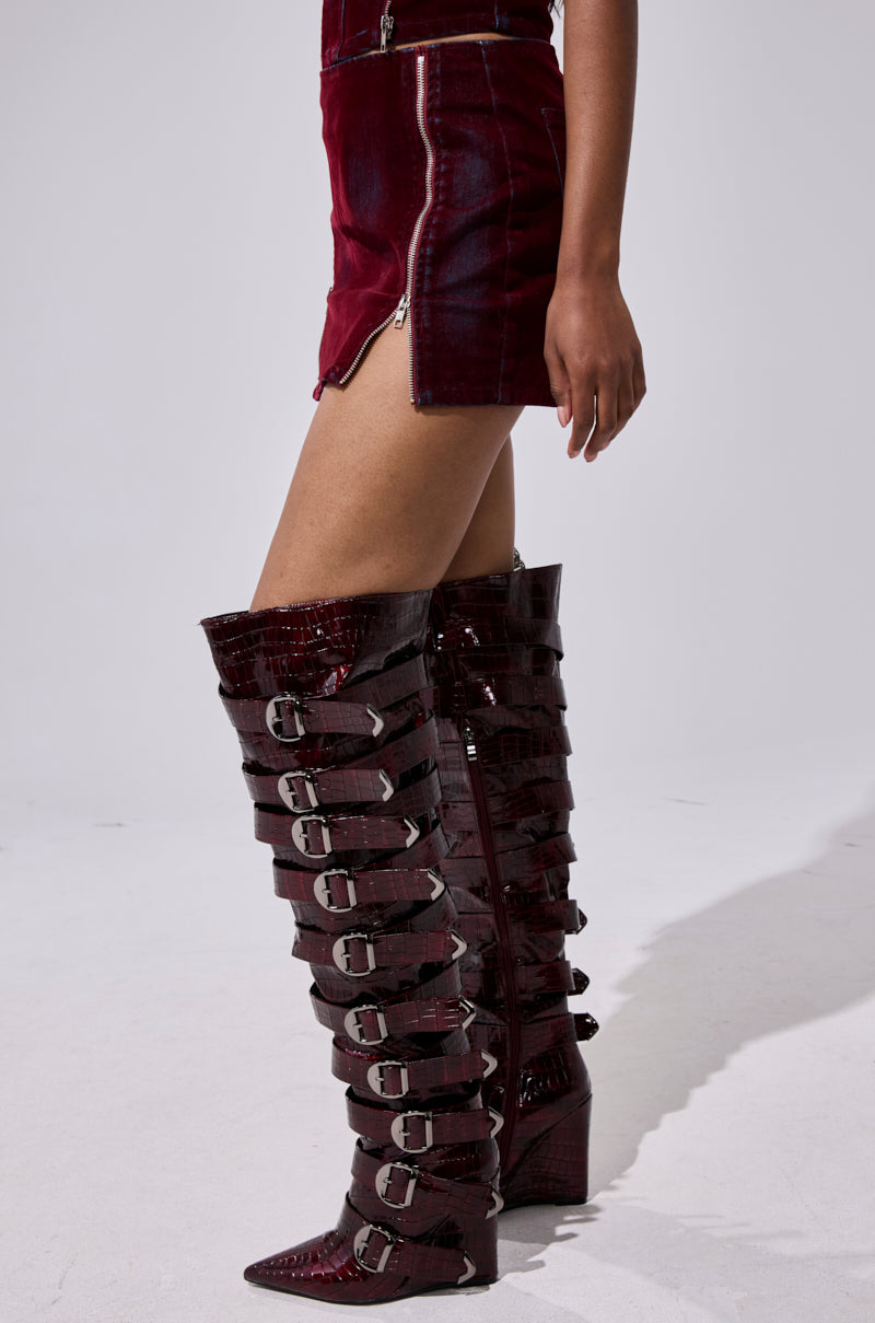 A person is wearing a shiny dark red FOLD OVER ZIP MINI SKIRT and matching knee-high boots with multiple silver buckle straps, standing against a plain light background. 5