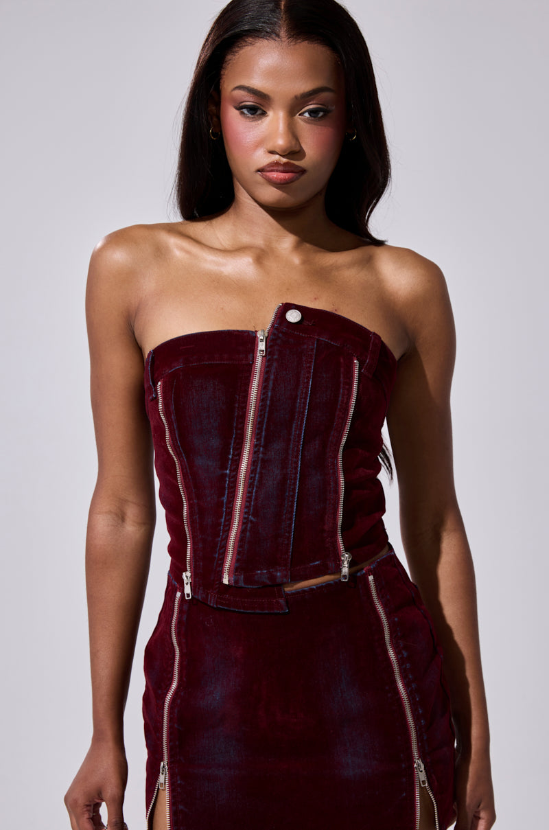 A woman wears the ASYMMETRIC ZIP CORSET in rich burgundy velvet with zippers and a matching skirt, standing against a plain light gray background. 0