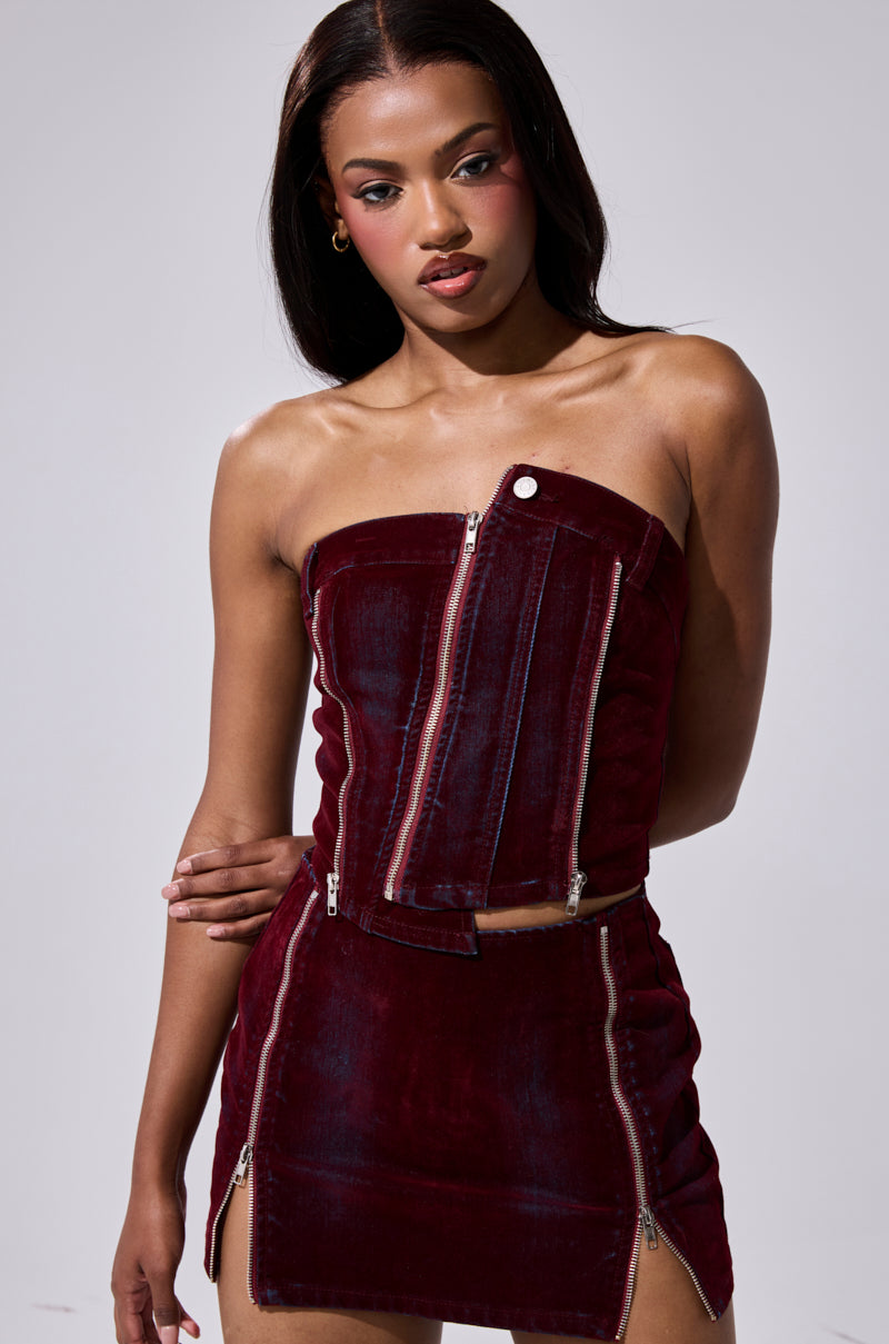 A woman poses confidently against a plain light background in the ASYMMETRIC ZIP CORSET—a strapless, deep red velvet top with asymmetrical zippers—paired with a matching high-waisted mini skirt featuring zipper details. 1