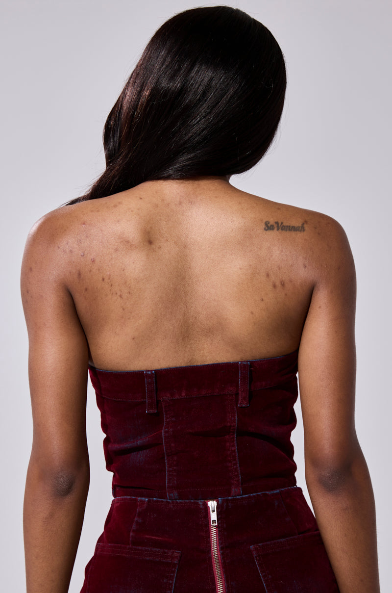 A woman with long dark hair is seen from behind wearing the ASYMMETRIC ZIP CORSET in burgundy denim, revealing natural skin texture and a "So Vannah" tattoo on her shoulder. 3